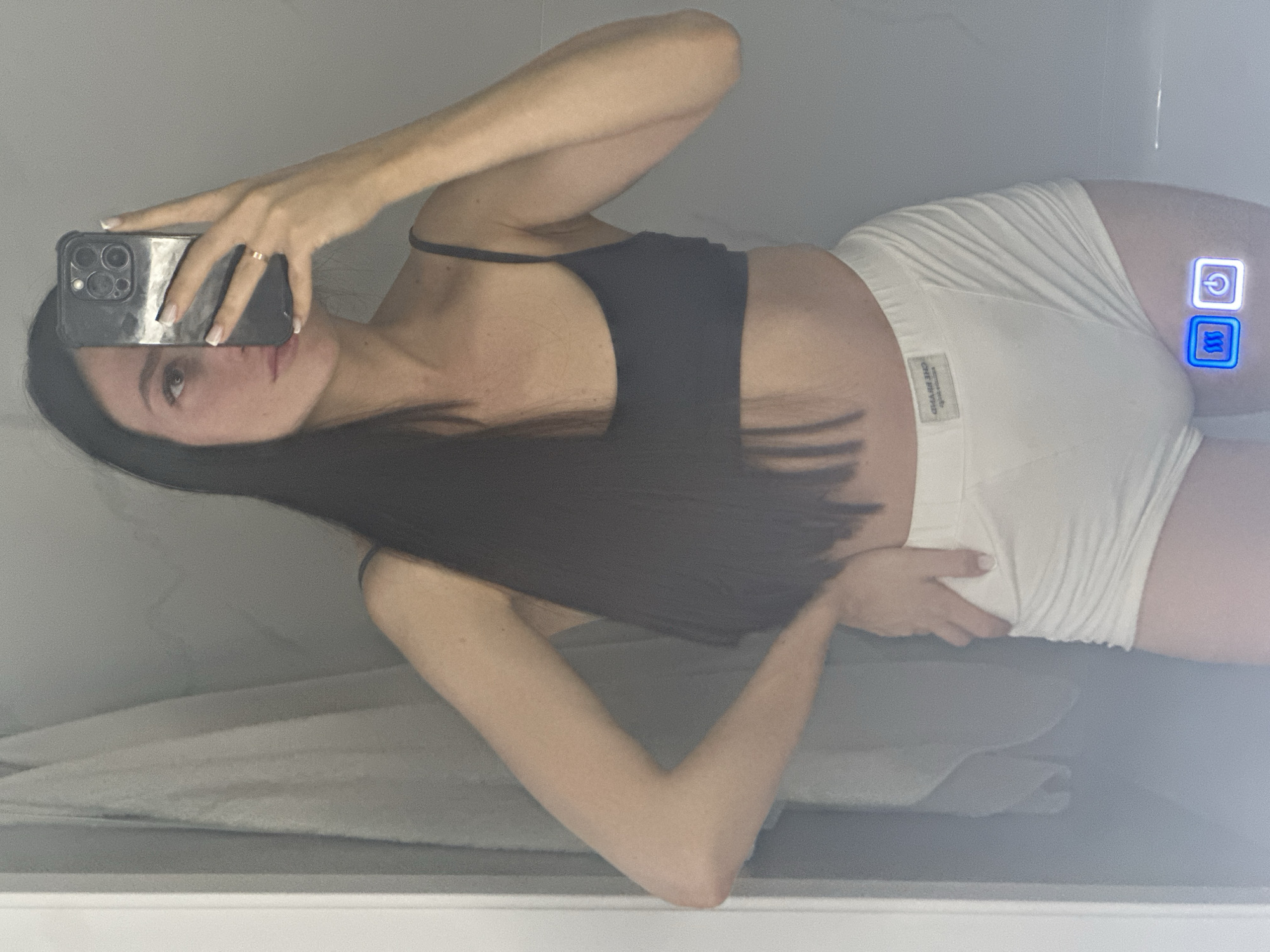 A woman with long dark hair and a fair complexion is taking a mirror selfie in what appears to be a bathroom. She is wearing a dark bra and white underwear. Her left hand is on her hip, and she is holding a smartphone in her right hand, angled to capture her reflection. The background is a tiled wall and a towel hanging on a rack. There are two illuminated buttons visible on the lower right side of the mirror, one with a wave icon and the other with a power icon. The image does not provide any information about the location being in Эстачи, Russia.