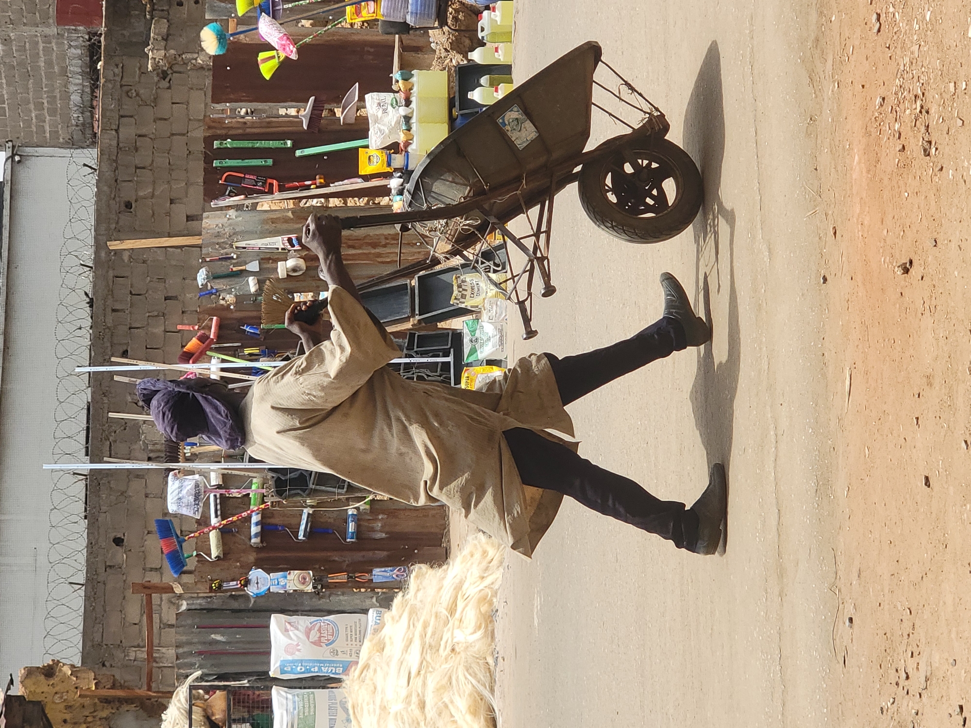 A man is depicted in an outdoor scene, pushing a wheelbarrow on a dusty path or street. He wears a light-colored tunic or robe over dark trousers and dark shoes, with a dark head covering. His hands are gripping the handles of the wheelbarrow, which features a single wheel and appears somewhat modified.
The background reveals what appears to be a street vendor's stall or market display. Various household goods and tools are arranged on shelves and boards against a wall made of cinder blocks and corrugated metal. These items include brooms, brushes, hand saws, paint rollers, small containers, and bottles of liquid. White plastic bags are visible, one clearly labeled "PLASTIC" and "BUA P.O.P," and another with "EXPRESS POWDER." A yellow bottle also displays the word "ROYAL." Barbed wire fencing can be seen along the left side of the background.
In the foreground, a large pile of light-colored, fibrous material, possibly jute or raw cotton, rests on the ground. The bright sunlight casts long shadows from the man and the wheelbarrow, suggesting it is either mid-morning or late afternoon on a clear, sunny day. The setting implies a commercial area in Maiburiji, Nigeria, with everyday activities taking place.