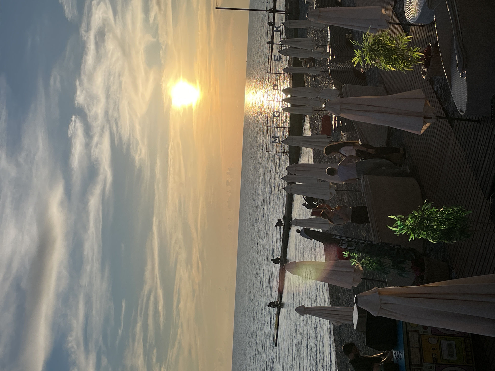 The image depicts a sunset scene at a beach in Sochi, Russia. The sun is setting over the calm, dark water of the Black Sea, casting a golden reflection on the surface. In the foreground, a wooden deck is laid out with numerous closed beach umbrellas and lounge chairs. Several people are visible on the beach and on a pier extending into the sea. Some are interacting, while others appear to be relaxing or working. A sign with Cyrillic letters, "МОКРР", is visible on a structure by the water. The sky is filled with wispy clouds, illuminated by the warm hues of the setting sun. The overall atmosphere is tranquil and serene, characteristic of a beach at dusk.