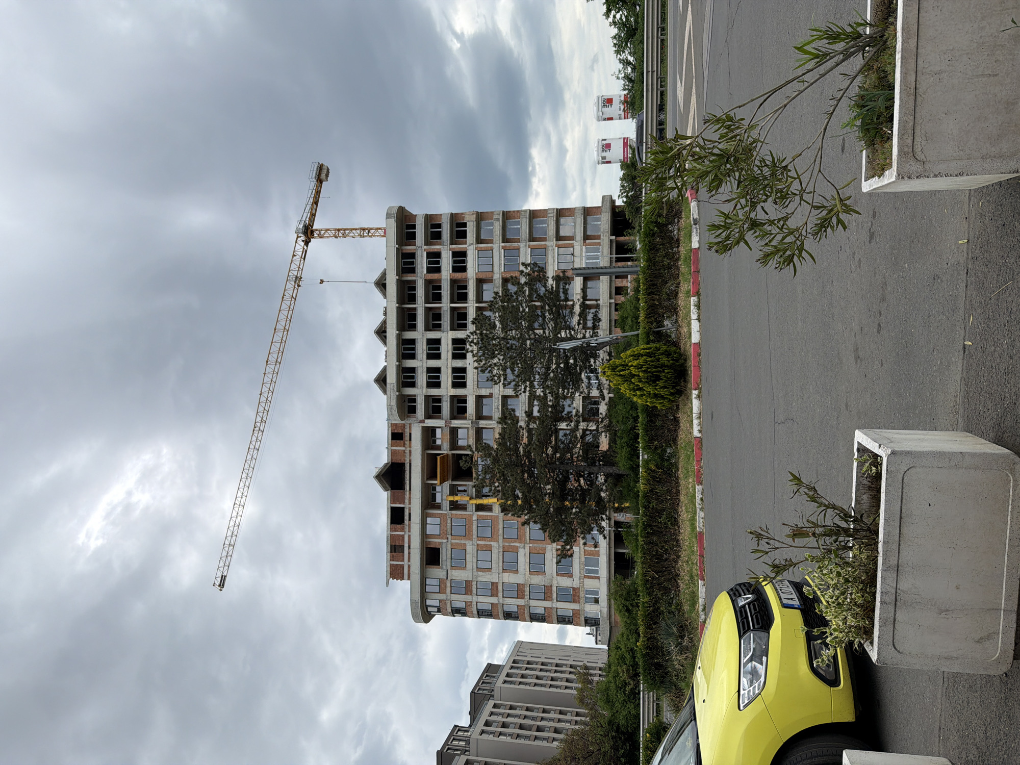 The image depicts a construction site in Burgas, Bulgaria. A high-rise building is under construction, with a crane present at the site. The building has multiple floors, and the exterior construction is visibly incomplete. A yellow car is parked near a concrete planter in the foreground. The sky is overcast with dark clouds. There are sidewalks and roads visible, along with landscaping including shrubs and trees. Other buildings are seen in the background. A street with a red and white curb borders the construction site. Advertisements for "MIT" are on top of another building in the background.