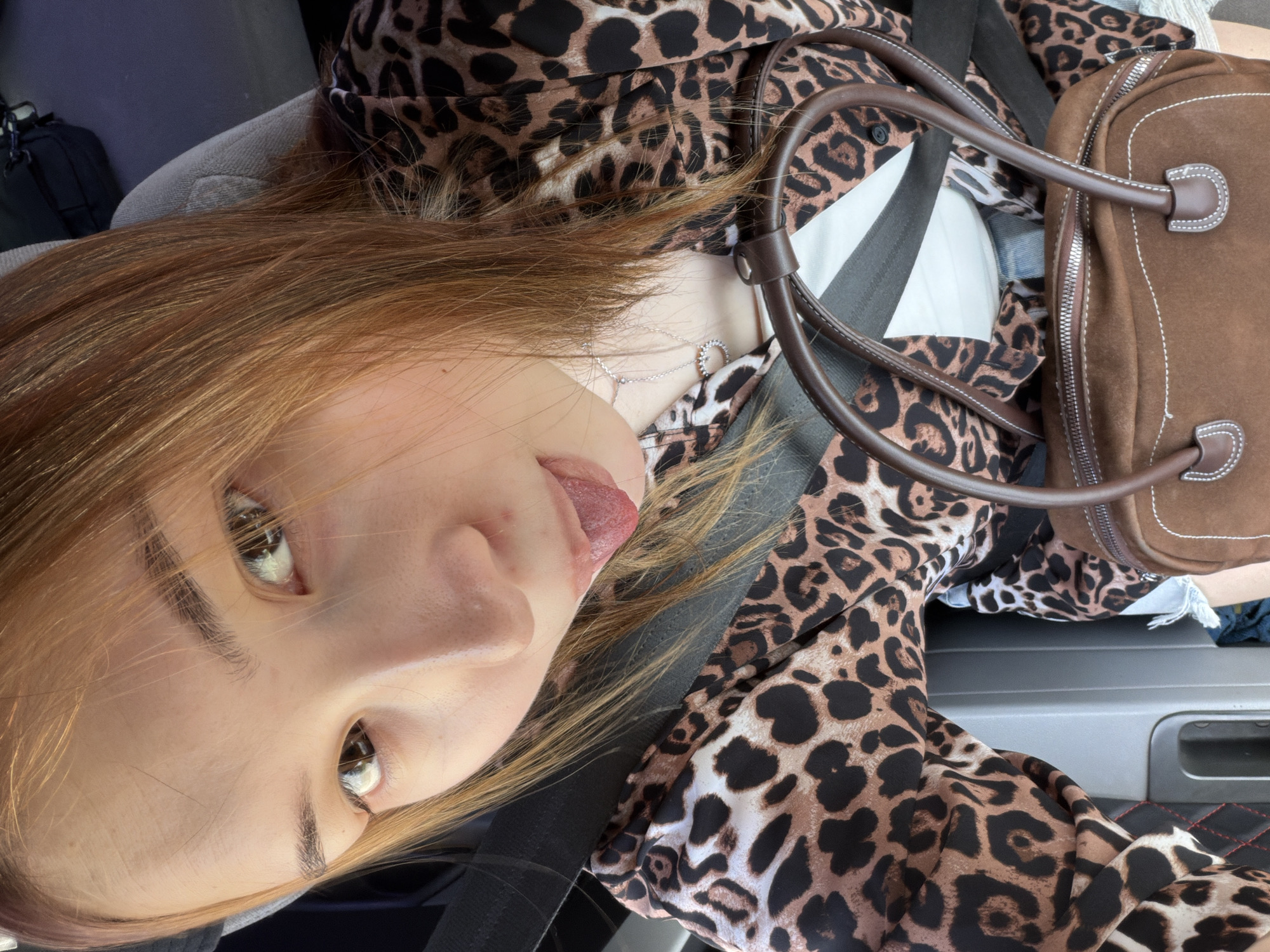 The image shows a person with auburn hair wearing a black seatbelt and a leopard-print top. The person is sticking out their tongue. A brown leather bag is visible near their right shoulder. The setting appears to be inside a vehicle. There is no explicit indication that the location is Chaikovsky, Russia.
