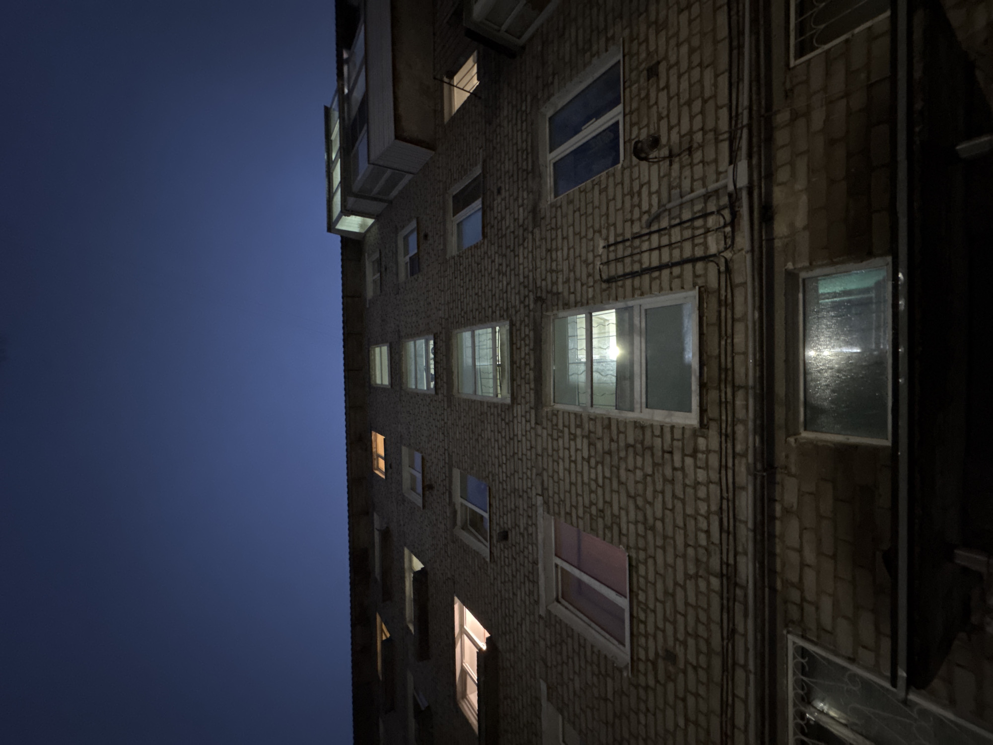 An upward shot shows a brick building at night with a blue sky. The building has several windows; some are illuminated, and some are dark. There are pipes running along the outside of the building. A balcony or structure is attached to the building's left side. The location is Chaikovsky, Russia.