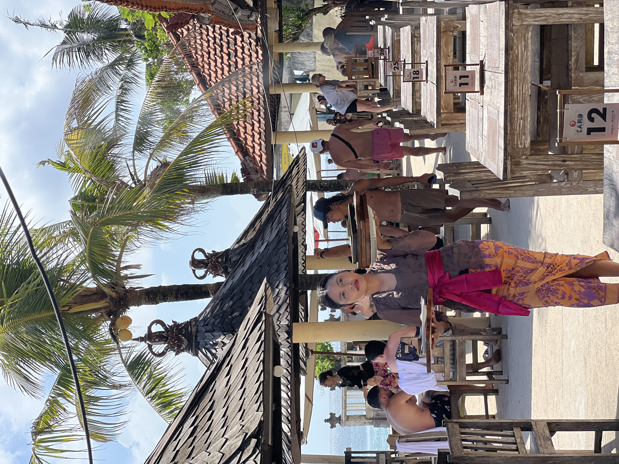 The image shows a partially obscured woman in traditional Balinese attire seated at a wooden table. In the background are multiple tables, each marked with a number (11, 12, 18, 25). People are seated or standing near the tables. Palm trees and a traditional Balinese-style roofline are also visible, suggesting a seaside restaurant in Kuta Selatan, Indonesia.