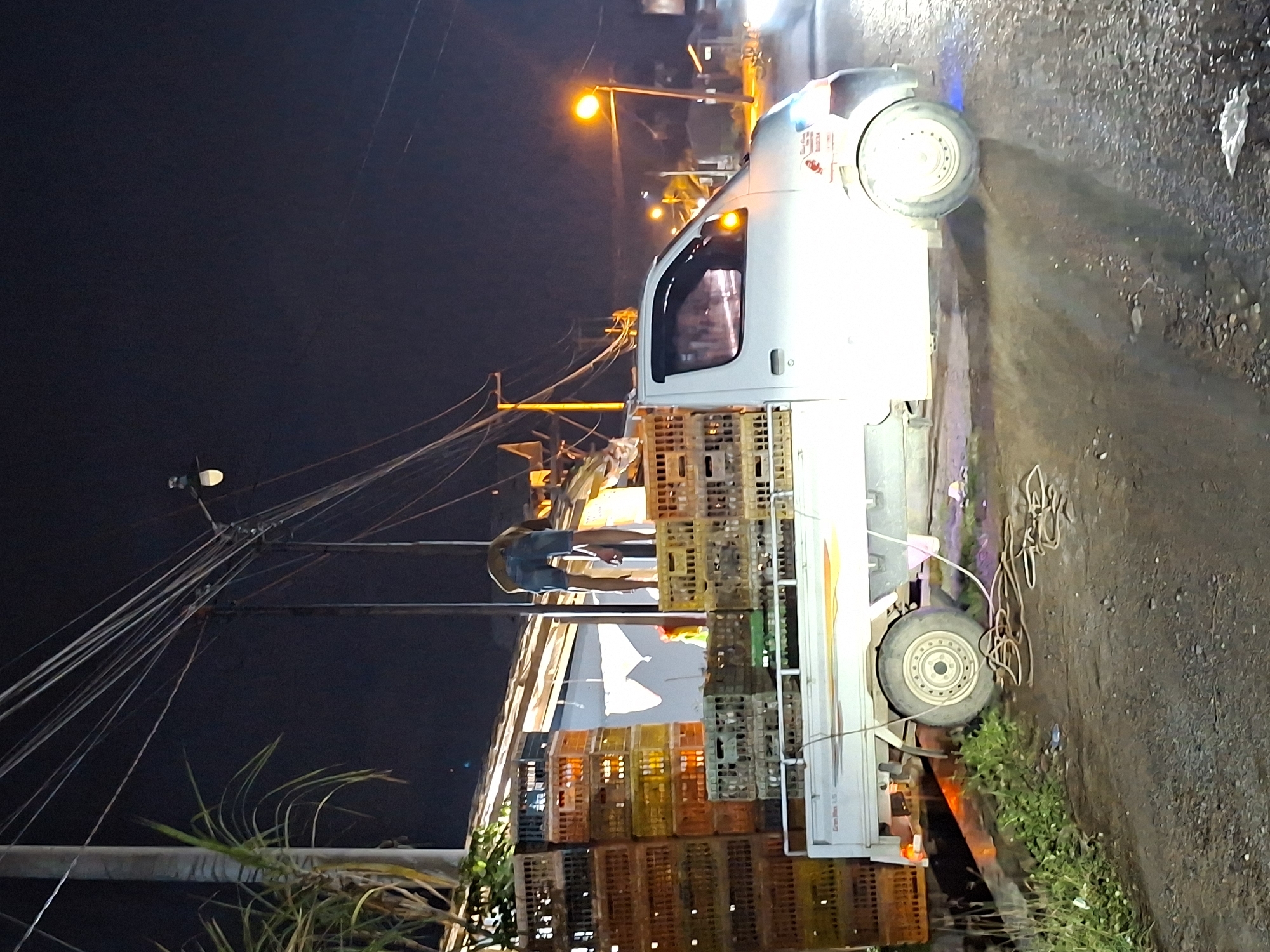At night in Lhokseumawe, Indonesia, a white pickup truck is heavily laden with stacked crates. A man stands atop the crates. Streetlights illuminate the scene. Power lines run above the scene. The road is wet. Vegetation is present on the side of the road.