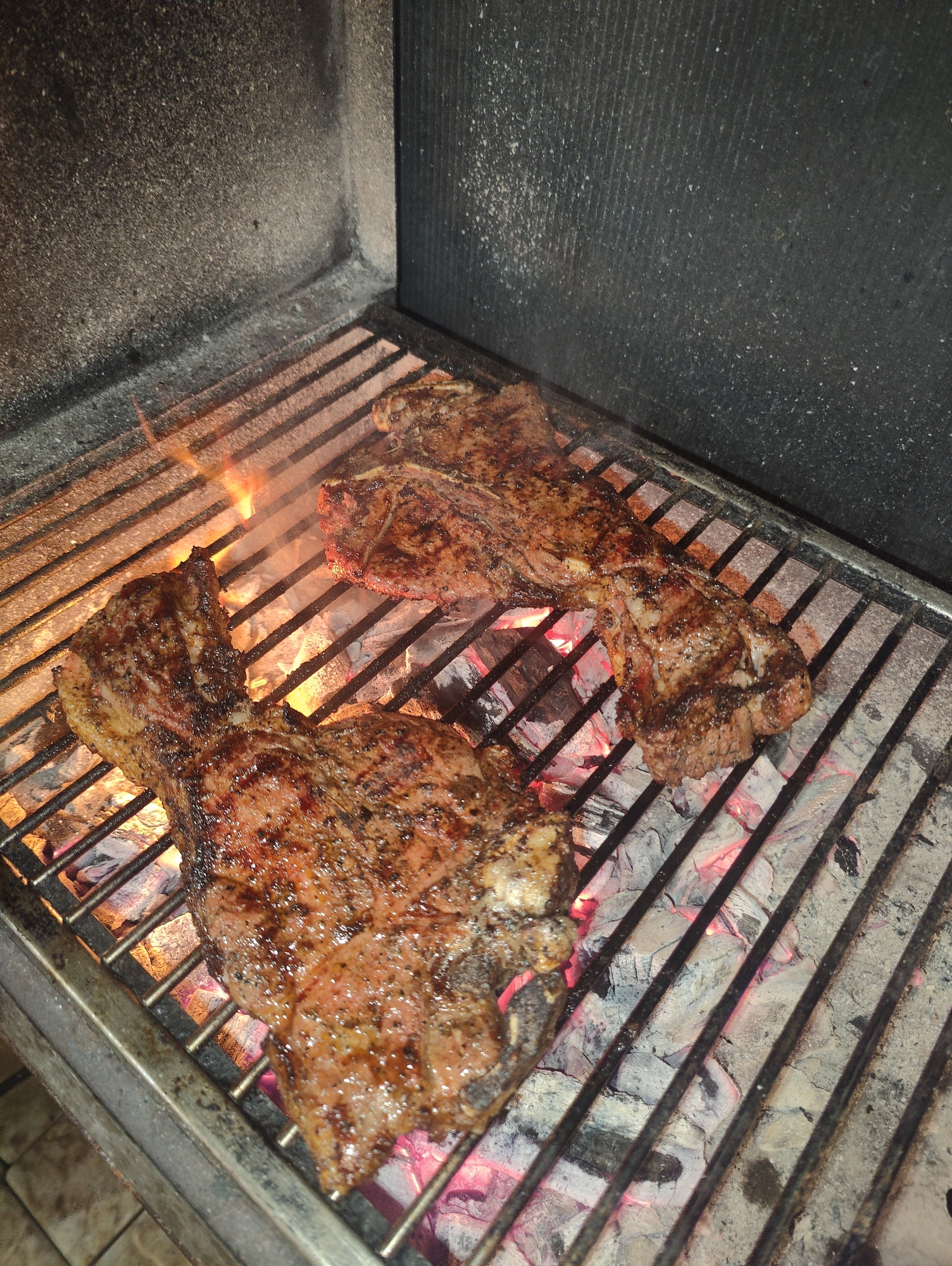 The image shows three steaks grilling over hot coals inside a grill. The steaks appear to be well-seasoned with visible grill marks. Flames are present, indicating the cooking process is active. The environment appears to be outdoors, possibly a backyard or a dedicated grilling area. There is no indication of the city of Izberbash, Russia, in the image or its context.