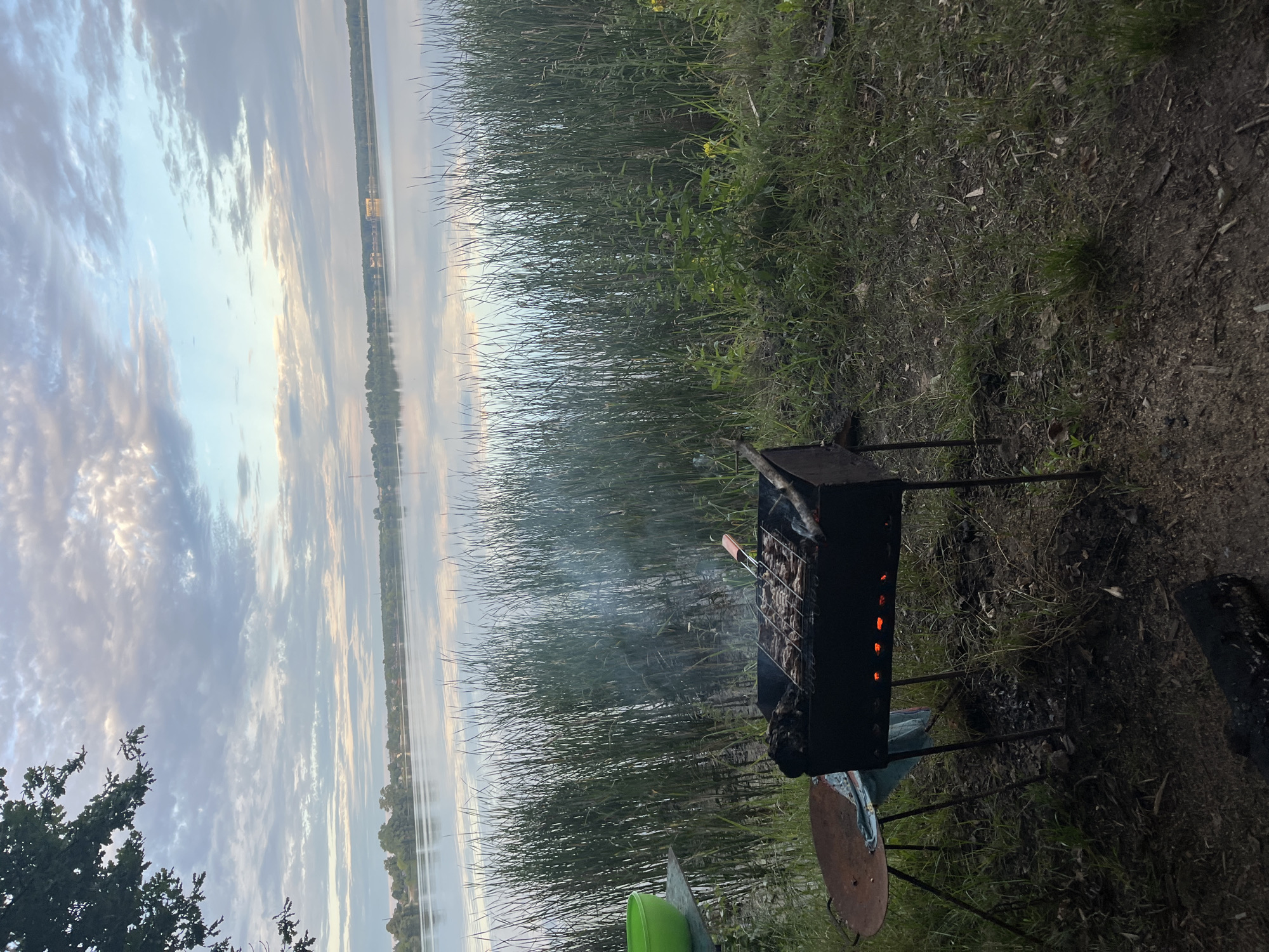 The image depicts an outdoor scene by a body of water, likely a lake or large river, in the vicinity of Вяззеўскі сельскі Савет, Belarus. The water is calm and reflects the cloudy sky, which features a mix of grey and pale blue hues with wispy clouds. Reeds and tall grass line the edge of the water, obscuring the background which appears to be a treeline and some structures in the distance.
In the foreground, a metal barbecue grill is set up on the grassy bank. Meat is cooking on the grill, with smoke rising from it. The coals beneath are glowing red. To the left of the grill, a rusty, circular metal surface on legs, possibly a makeshift table, is visible. A green plastic bowl rests on a small table or shelf to the left of that. The ground is covered in grass and dirt, with some fallen leaves and debris. A charred log lies on the ground in the lower foreground. The overall impression is that of a casual outdoor meal being prepared by the water. No people are visible in the frame.