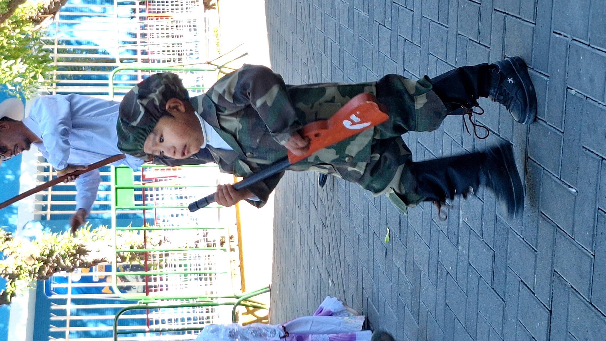 A young child in a camouflage military costume stands on a gray brick sidewalk in Cerro Colorado, Peru. The child holds a toy rifle with an orange stock. Another person, possibly an adult, is visible in the background wearing a white shirt and hat while holding a long stick or pole. A green and white fence is also visible in the background. Trash or discarded items are on the sidewalk near the child.