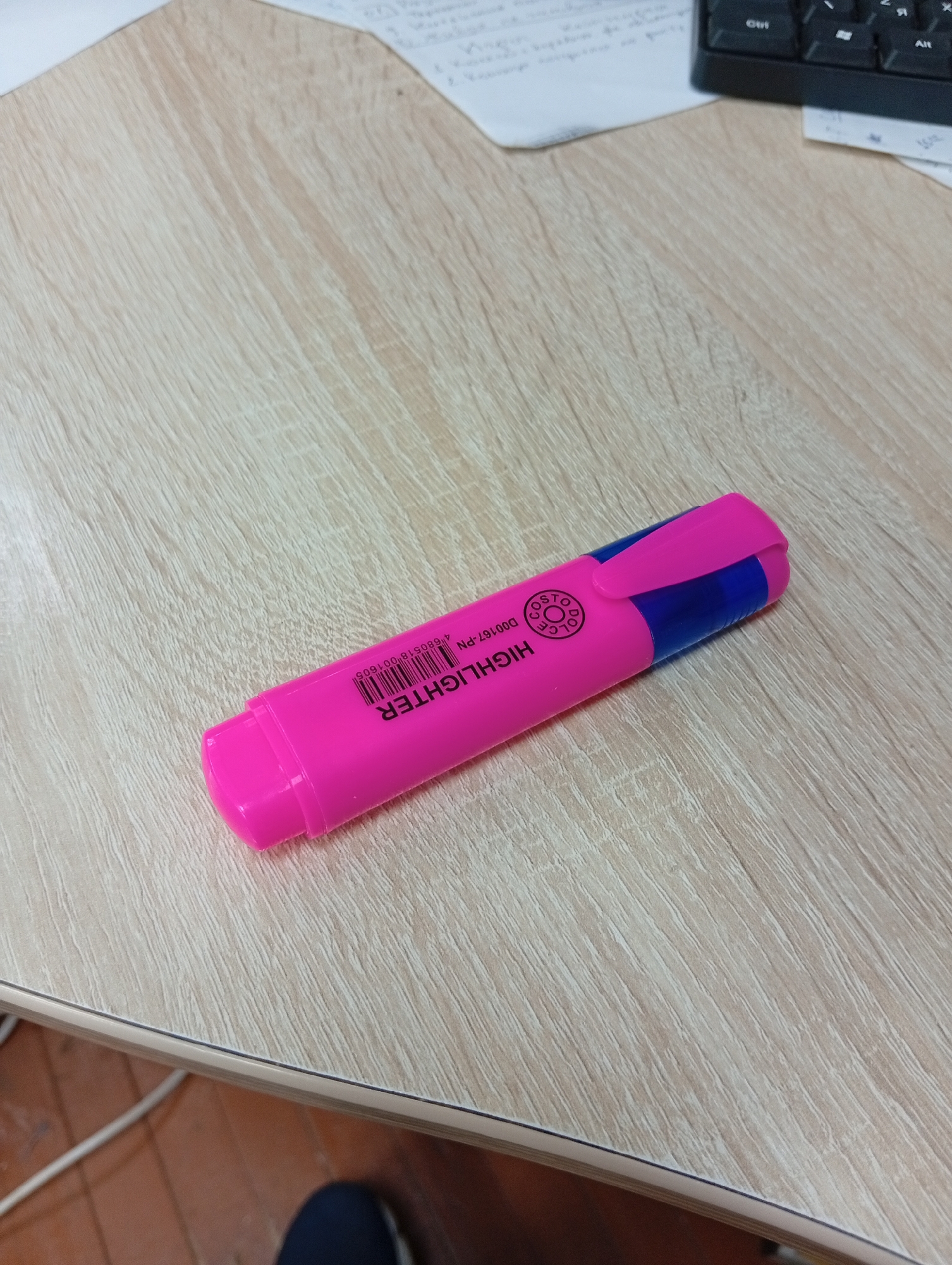 A bright pink highlighter with blue accents lies diagonally on a light wood grain desk. The highlighter has black text printed on it, including a barcode and the word "HIGHLIGHTER". Part of a keyboard and some papers are visible in the upper right corner of the image. The image was taken in Крыўлянскі сельскі Савет, Belarus.