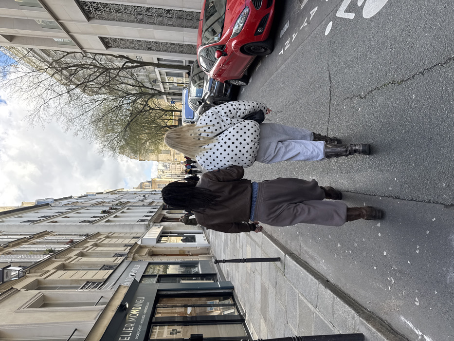 An outdoor, eye-level street view captures two individuals walking away from the camera on a paved road in Paris, France, identified by the visible "ATELIER MONDINEU" sign at 12 Rue de Longueville. The person on the left wears a brown bomber jacket, dark brown wide-leg trousers, and brown boots, with dark hair cascading down their back. The person on the right has blonde hair, a white puffer jacket with black polka dots, light grey sweatpants, and dark chunky boots, with a black shoulder bag worn cross-body. This individual holds a lit cigarette in their right hand.
To the left, a stone sidewalk runs alongside multi-story Parisian buildings housing businesses, including "ATELIER MONDINEU" and "ES. LONGUEVILLE". Two black bollards are positioned on the sidewalk. To the right, a row of vehicles is parked along the curb, prominently featuring a red Ford Fiesta in the foreground, followed by a dark SUV. The street itself is asphalt with visible cracks and white painted markings, including a bicycle symbol and directional arrows. Buildings line both sides of the street, with older, traditional architecture on the left and a more contemporary structure with vertical grilles on the immediate right. Bare trees are visible further down the street. The sky is overcast, providing diffuse daylight.