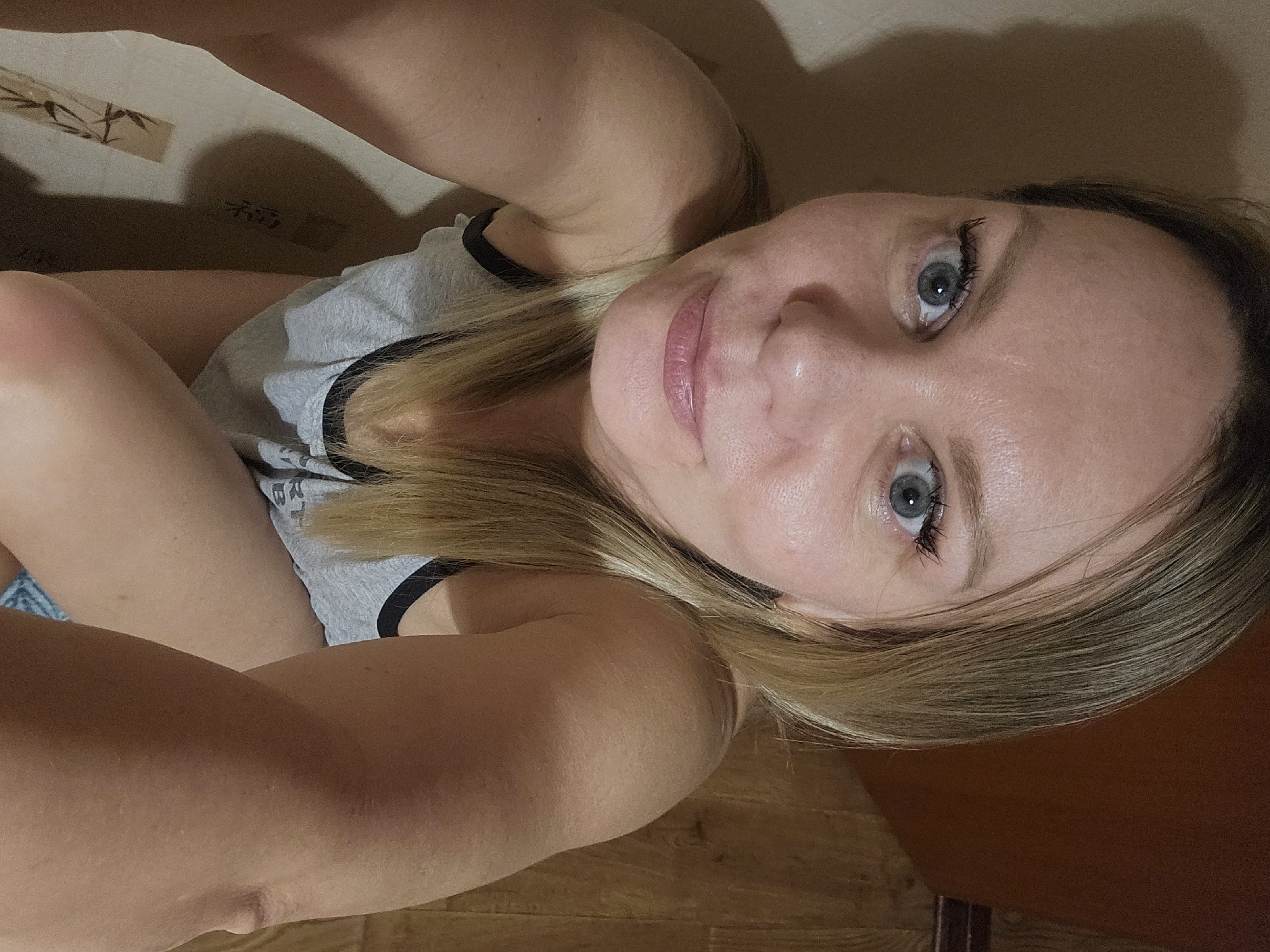 A blonde woman with fair skin and blue eyes is looking upwards into the camera. She is wearing a light gray tank top with black trim around the neck and armholes. She is inside a box with a beige interior, which appears to have a bamboo design. The background consists of a light brown hardwood floor and a dark brown wooden cabinet. The location is unknown and no evidence suggests it is in Sevastopol, Ukraine.