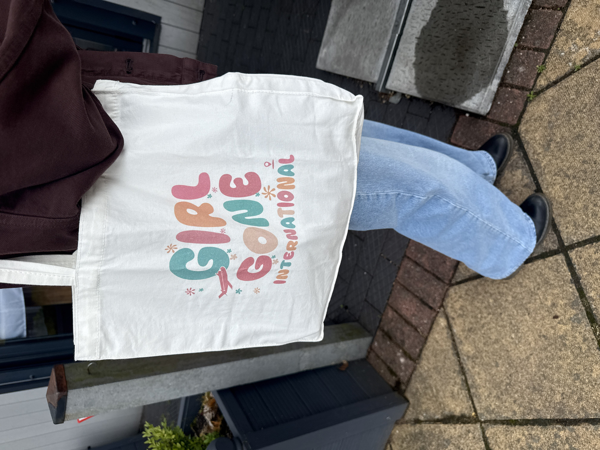 An individual stands outdoors, partially visible from the mid-torso downwards. They wear a dark reddish-brown long-sleeved garment, light blue wide-leg jeans, and black closed-toe shoes. The person holds a white canvas tote bag with their left hand. The bag features a colorful graphic design with the text "GIRL GONE INTERNATIONAL." "GIRL" and "GONE" are rendered in large, bubbly, multi-colored letters (teal, orange, pink), while "INTERNATIONAL" is in smaller orange letters. Small pink airplanes, various asterisk-like symbols, and a pink location pin icon are also part of the design. The ground is paved with a mix of tan/light brown concrete pavers, reddish-brown brick pavers, and dark gray/black rectangular pavers. To the left, a dark gray planter box with green foliage and a gray concrete post with a dark brown cap are visible. To the right, a dark building structure and a dark gray metal grate on the ground with a wet appearance are present. The scene takes place in the city of Unknown, Unknown.