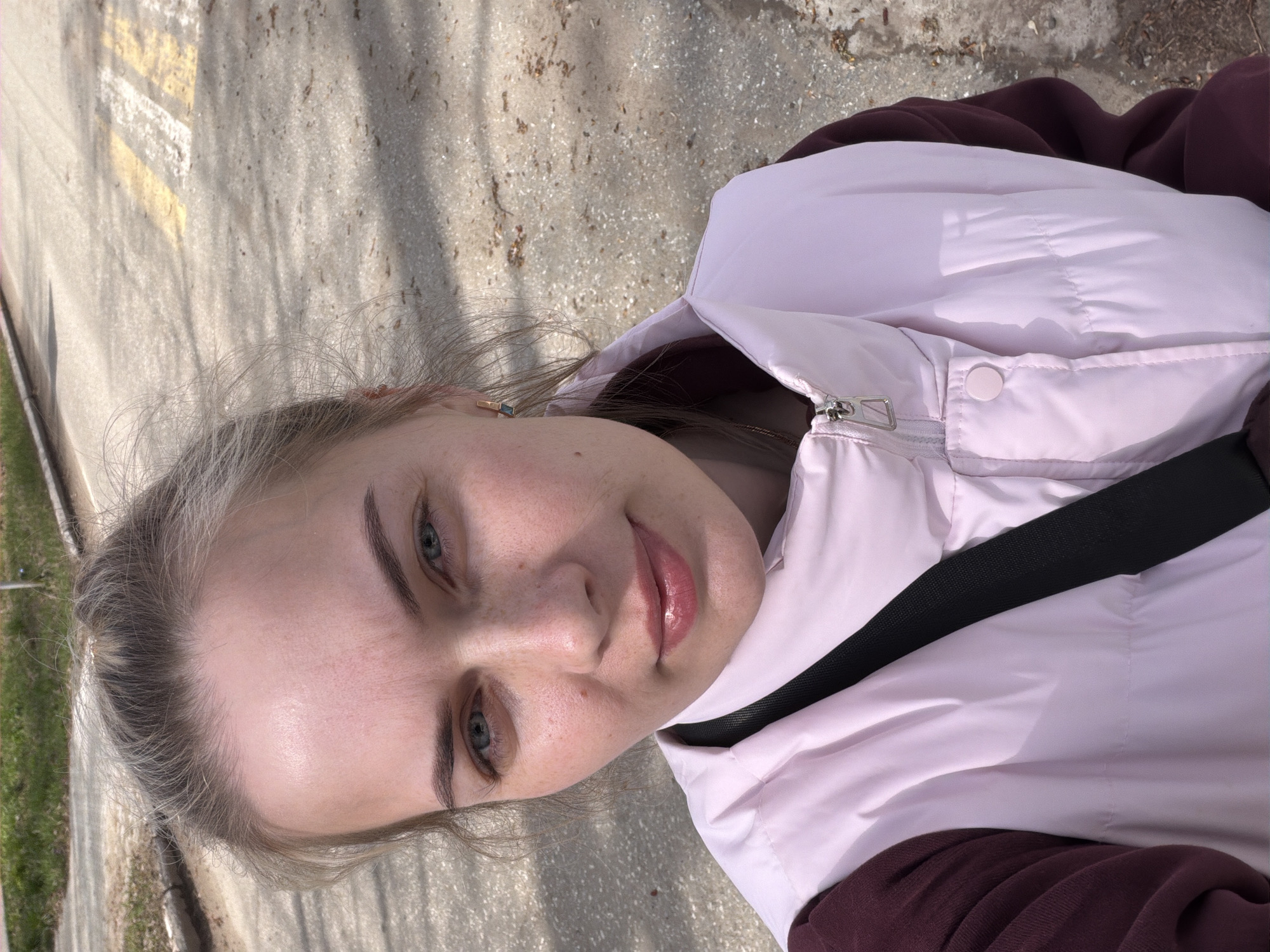 The media file features a close-up, slightly tilted selfie of a young woman taken outdoors in Chaikovsky, Russia. The woman, with fair skin, light blonde hair pulled back, and light-colored eyes, looks directly at the camera with a subtle, pleasant smile. Her lips have a glossy pink tint. She has visible freckles across her nose and cheeks and neatly shaped eyebrows. She is wearing a light pink puffer vest over a dark maroon long-sleeved top, with a black strap (possibly from a bag or seatbelt) crossing her chest. A small, square-shaped earring, possibly gold with a blue or green stone, is visible on her left ear.
The background consists of a paved surface, likely a road or pathway, with distinct yellow and white lines indicating road markings. The ground shows varying textures and shades, with shadows from overhead trees or structures dappling the surface, suggesting a sunny day. Green grass and some dirt are visible along the left edge of the frame. The overall impression is a casual outdoor portrait on a bright day. There is no visible text in the image.