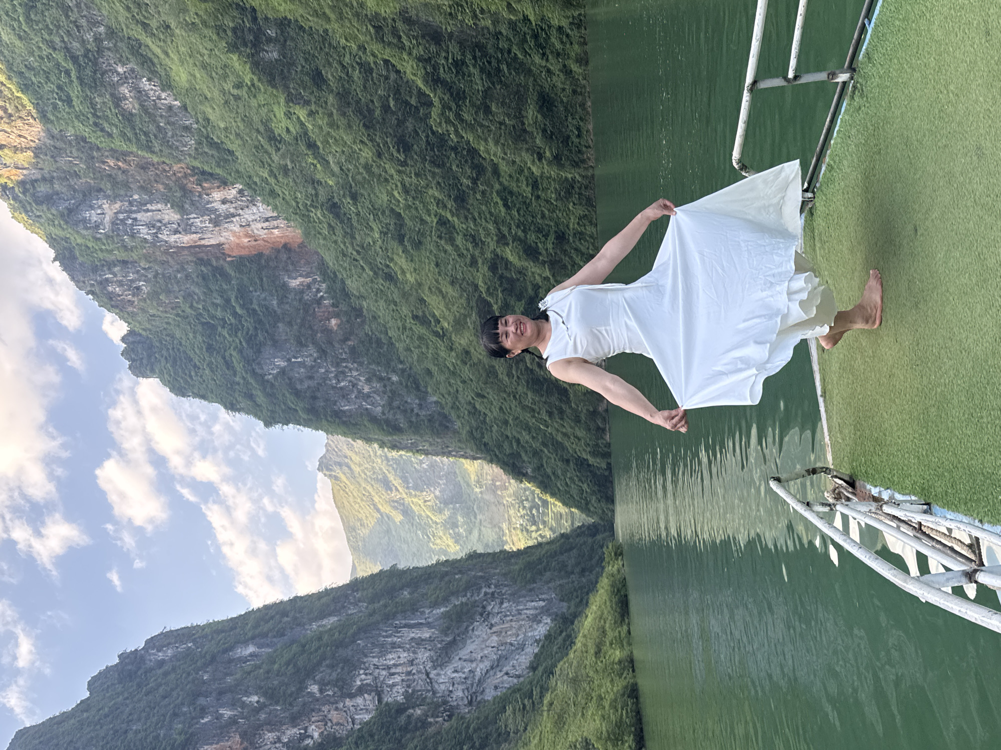 A woman in a white dress stands barefoot on a green, artificial-turfed platform with a white railing, holding the edges of her dress and smiling. The platform appears to be the deck of a boat on a river or lake, as water surrounds it. Towering, tree-covered mountains rise steeply on both sides of the water. The sky is partly cloudy, with sunlight illuminating the landscape. This scene is likely located in Đồng Văn Commune, Vietnam, known for its dramatic karst landscapes.