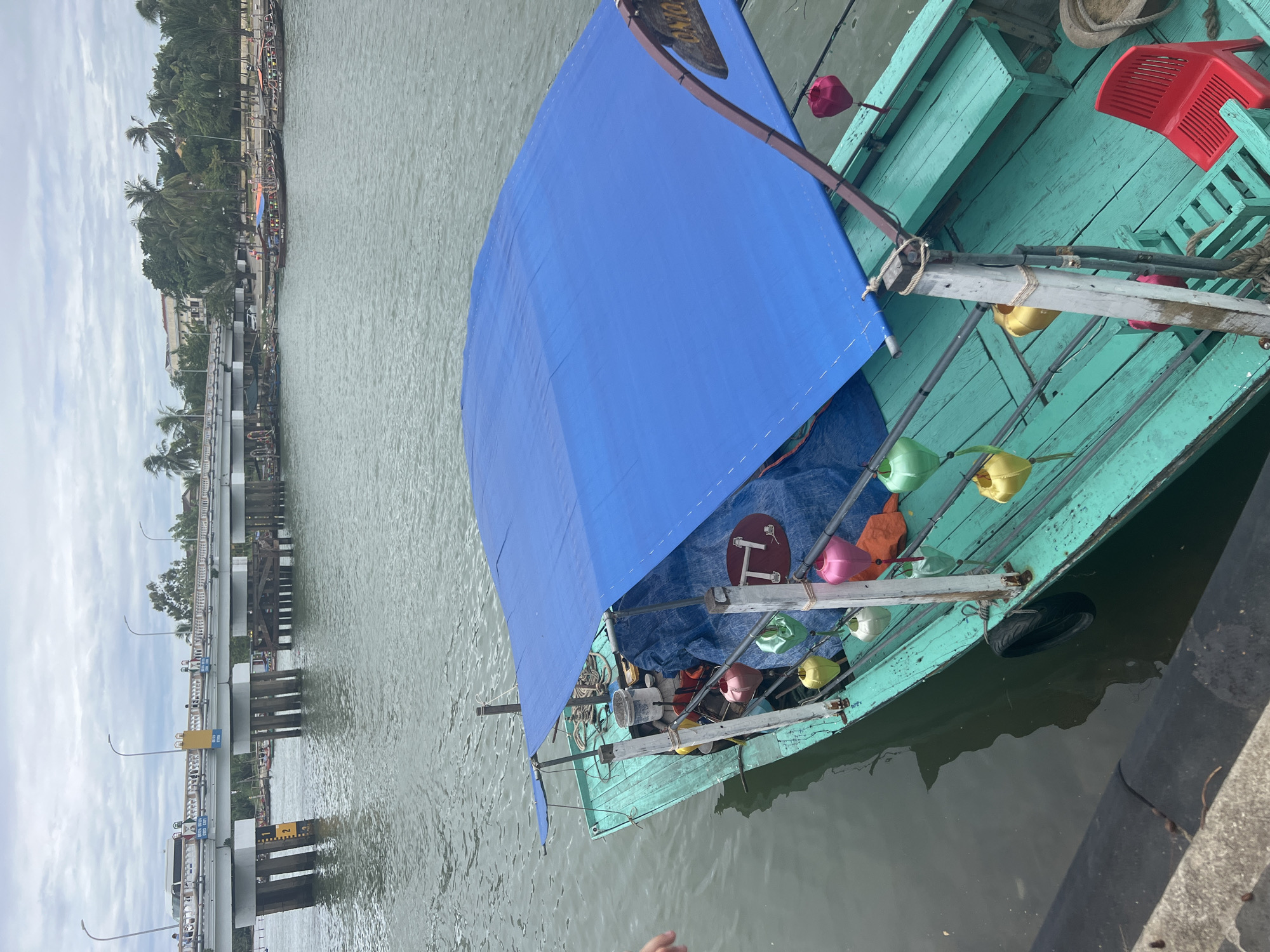 A boat is docked in a river in Hoi An, Vietnam. The boat is teal and has a blue tarp covering its top. Colorful lanterns hang from the sides of the boat. In the background, a bridge spans the river, with buildings and palm trees lining the shore. The sky is overcast.
