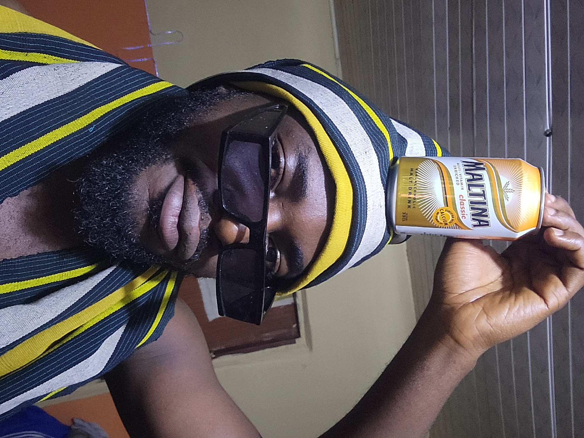 A man wearing a striped beanie and dark sunglasses balances a can of Maltina on his head. He has a beard and is wearing a striped scarf. The background shows a ceiling and a wall with a doorway. The image is a selfie taken indoors, likely in a residential setting in Jos, Nigeria, given the subject's attire which is reminiscent of traditional Nigerian clothing.