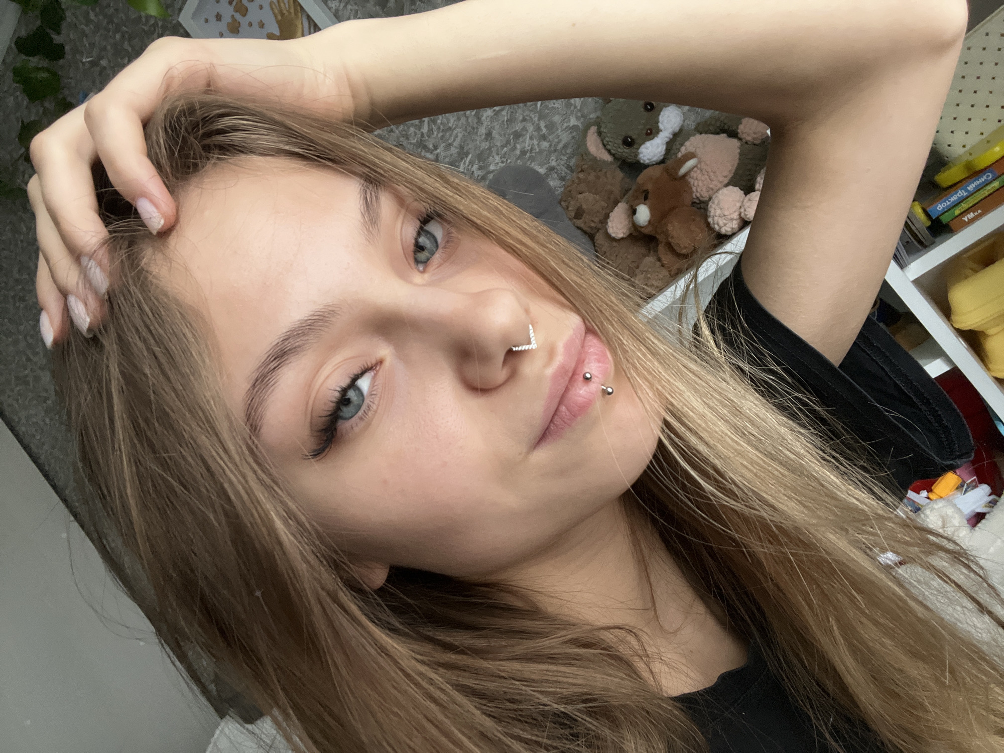 A fair-skinned young woman with light brown hair and blue eyes is captured in a close-up, angled selfie. She has a silver V-shaped nose piercing and a lip piercing on the left side. Her right hand is raised, fingers touching her hair. She is wearing a black shirt. The background includes a gray carpet, a collection of stuffed animals, and a shelf with books and a yellow object. There is no indication of the location in the city of Фоки, Russia.