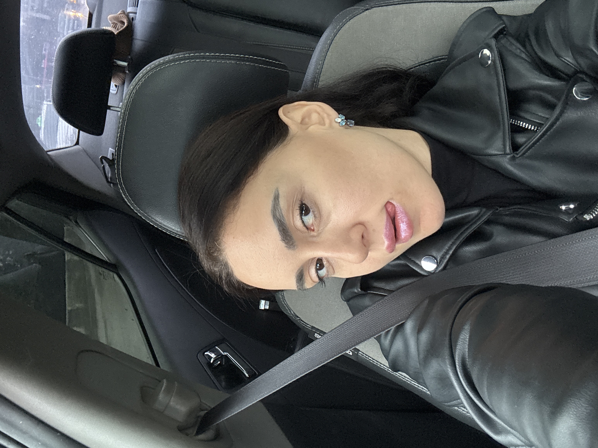 An adult female is depicted in the front passenger seat of a vehicle in Tbilisi, Georgia. She is viewed from the chest up, looking directly towards the camera. Her dark hair is styled down. She wears a black leather jacket over a black turtleneck garment, and a dark seatbelt is fastened across her left shoulder and chest. A cluster earring with blue and clear stones is visible on her right ear. Her lips are coated with a pink, glossy substance, and her tongue is partially extended from her mouth. The car interior features black leather seating with gray fabric inserts and white stitching visible on the headrests. Chrome door handle components are present on the passenger-side door. Through the blurred rear window, an urban street scene with indistinct artificial lights, including a faint green light, indicates dusk or nighttime conditions, with possible precipitation or condensation on the glass. A brown knitted object rests on the vehicle's rear parcel shelf.
