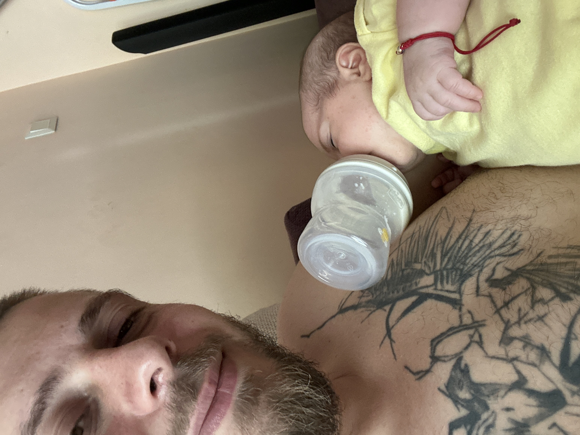 An overhead medium shot shows a man with a beard and tattoos on his chest feeding a baby from a bottle. The baby is wearing a yellow shirt and a red string bracelet on its wrist. The man is looking at the camera with a slight smile. The background is a plain wall with a light switch. The scene appears to be indoors, possibly a living room.