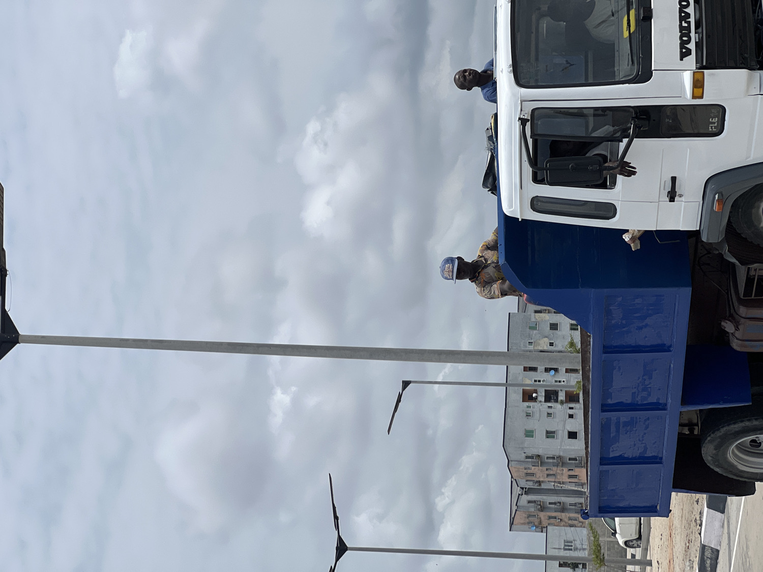 This full-shot, rotated image depicts an outdoor urban or semi-urban scene, featuring a large truck and several individuals under an overcast sky.
The main subject is a white and dark blue truck, possibly a dump truck or a heavy-duty flatbed, occupying the right side of the frame. The truck's cab is white, and its bed is a distinct dark blue. Visible on the front grille of the cab is the brand name "VOLVO" in white capital letters, along with the letters "FL" (likely part of a model designation) below it.
At least three people are associated with the truck. One man, wearing a blue shirt, is seen leaning out of the passenger side window of the cab, looking towards the viewer. Another man, dressed in a light-colored, patterned shirt and a blue baseball cap, is standing in the blue bed of the truck, looking downwards. A third person's hand is visible inside the cab, near the passenger door. They appear to be engaged in work-related activity, possibly inspecting or preparing the vehicle.
In the background, behind the truck, several multi-story buildings with numerous windows are visible, suggesting a residential or commercial area. Scattered streetlights or utility poles with horizontal arms are positioned on the left side of the frame, extending across the foreground and mid-ground.
The sky is largely covered with light grey and white clouds, indicating an overcast daytime. The overall lighting is diffused, with no harsh shadows, suggesting either early morning, late afternoon, or a heavily clouded day. The ground appears to be a paved surface.
The location context provided is Alaguntan, Nigeria.