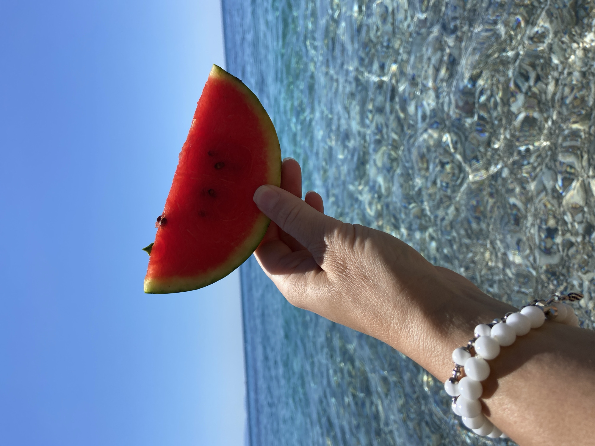 A hand holds a slice of watermelon in front of a bright blue sky and the Adriatic Sea. The watermelon slice is triangular with a green rind and bright red flesh speckled with seeds. The hand is adorned with a white beaded bracelet. The sea is a vibrant turquoise, with sunlight glinting off the pebbles visible beneath the clear water. The scene is set on a beach in Prijevor, Montenegro.