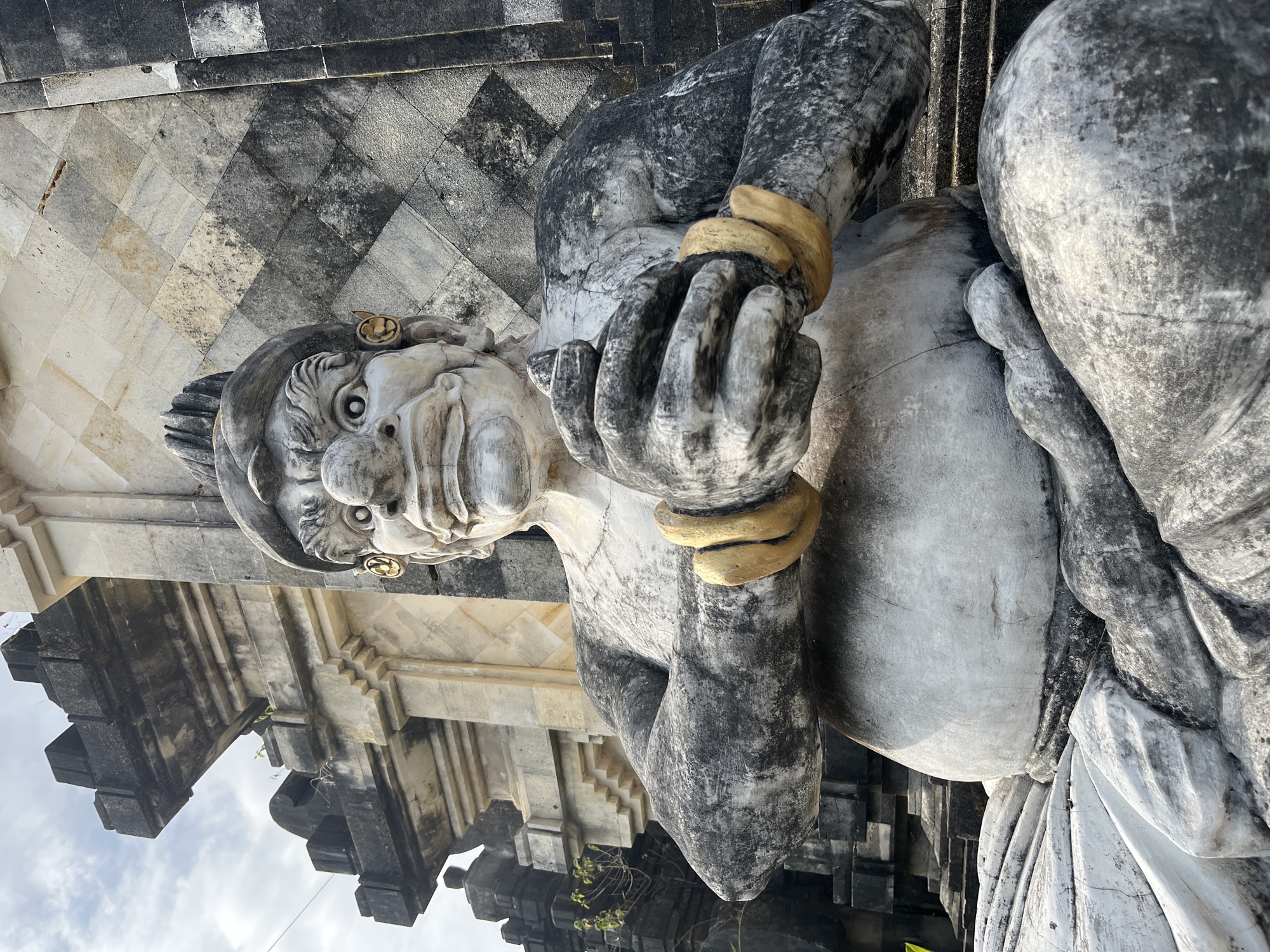 The image shows a stone statue of a creature with a large belly, large hands clasped together, and gold bracelets on its wrists. The statue is seated in front of a stone structure with intricate carvings and a checkered pattern on the wall. The sky is visible in the background. The statue is likely a guardian figure found at a temple or sacred site. This type of statue is commonly seen in Bali, Indonesia, and this particular one appears to be located in Kuta Selatan, a region known for its temples and cultural sites. The statue is weathered, showing signs of age and exposure to the elements. The lighting suggests it is daytime, with the sun casting shadows on the statue and the surrounding architecture.