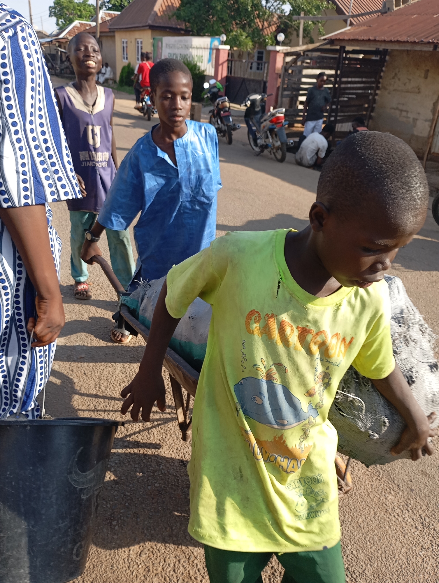 A group of boys are walking down a street in Shendam, Nigeria, carrying a cart loaded with two large sacks. A boy in a yellow t-shirt with a cartoon whale graphic is pulling the cart, while another boy in a blue tunic walks beside him. A third boy, wearing a purple tank top, walks ahead of them, looking back and smiling. The street is paved with gravel, and buildings with corrugated metal roofs line the background. In the distance, motorcycles and other vehicles can be seen. The lighting suggests it is daytime, likely late afternoon or early morning due to the long shadows.