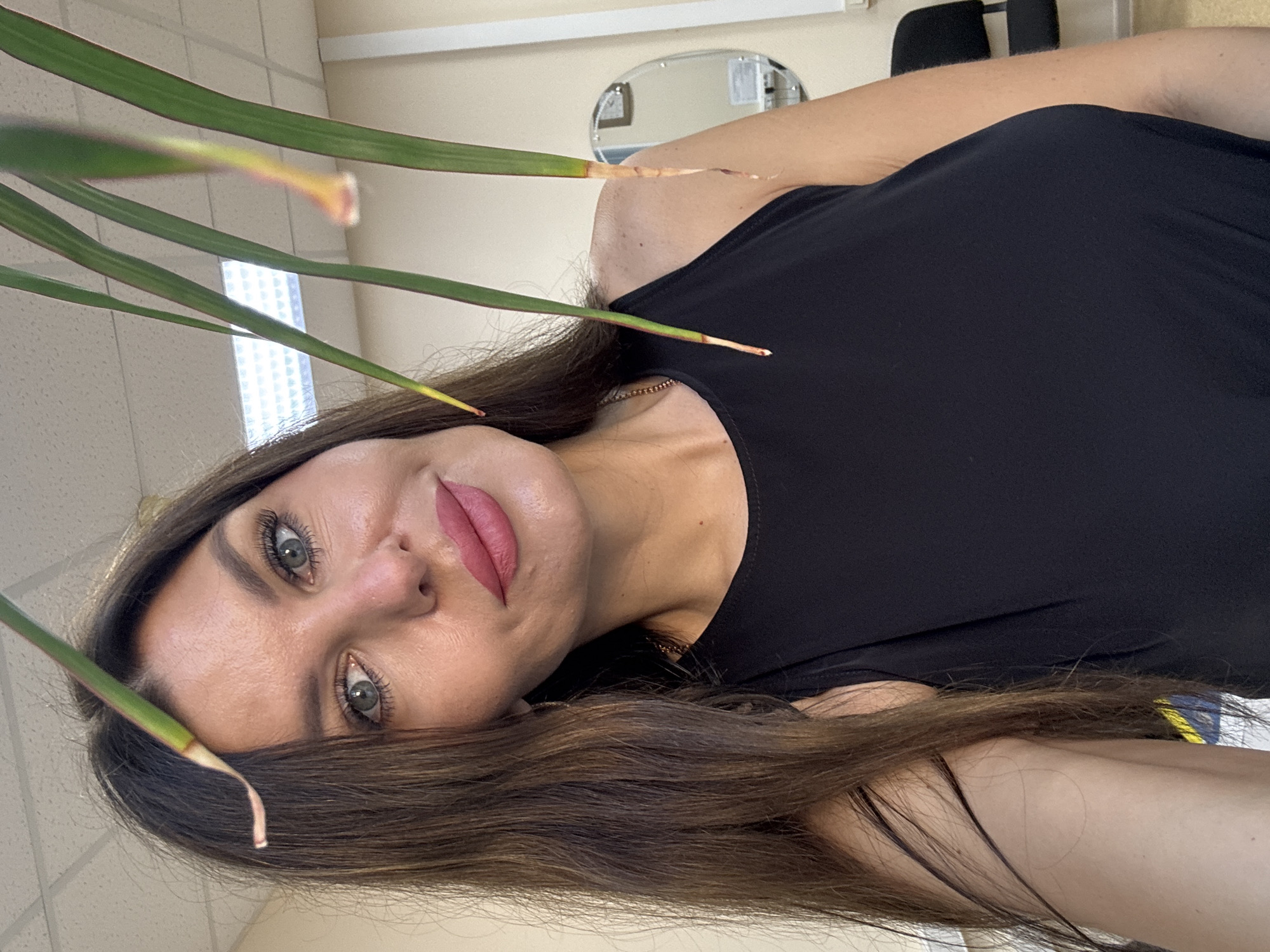A selfie of a woman in her 30s or 40s with long, dark brown hair and striking green eyes. She has fair skin with a light dusting of freckles, prominent cheekbones, and full, pink lips. Her makeup includes dramatic eyelashes and possibly subtle lip filler. She is wearing a black, sleeveless top. The background appears to be an indoor setting, possibly an office or a room with a suspended ceiling and fluorescent lighting. A plant with long, green leaves partially obscures the woman's face and shoulder. A circular mirror and a chair are visible in the background, suggesting a personal or professional space. The image quality is good, with clear focus on the woman. There are no discernible landmarks or indicators of the specific location within Chaikovsky, Russia, beyond the general indoor environment.