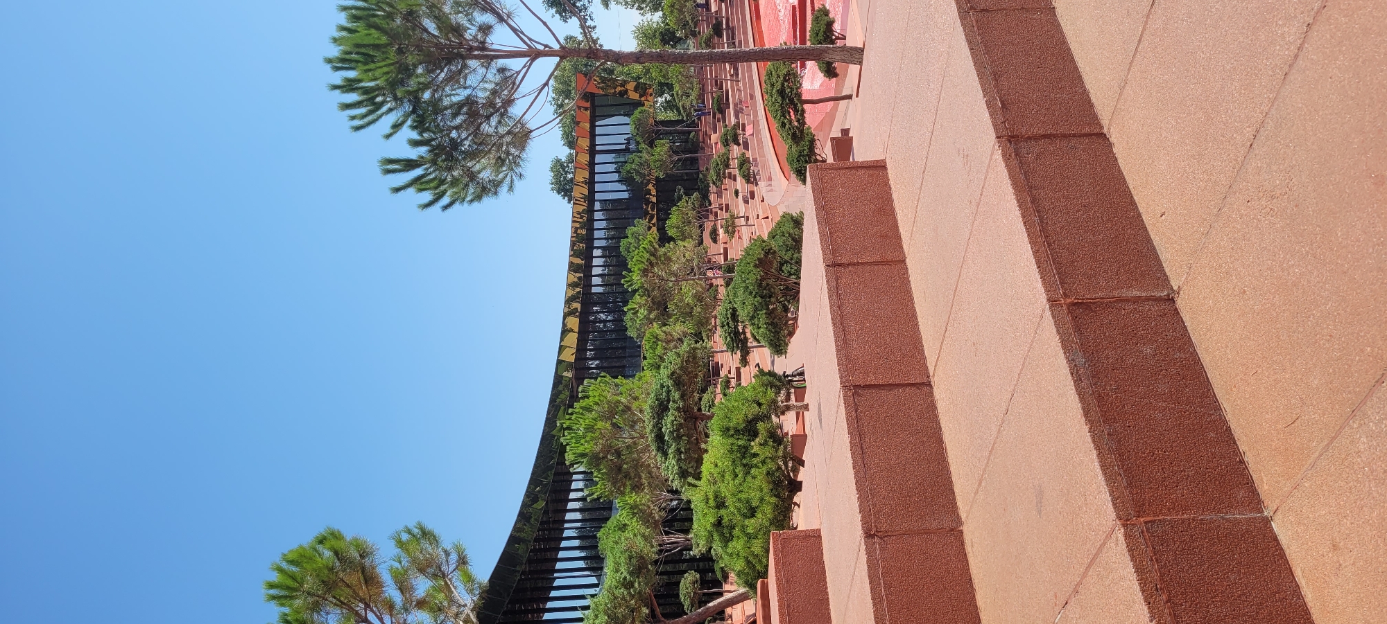 The image depicts an outdoor scene with a modern, curved building featuring a reflective, golden facade in the background. In the foreground, there are large, stepped structures made of reddish-brown material. The steps are bordered by a textured, concrete-like surface. A variety of carefully manicured trees, predominantly pine trees of varying sizes, are planted throughout the scene, some appearing to be in a Japanese garden style. The ground is paved with similar reddish-brown material as the steps. The sky is clear and blue, indicating a sunny day. Several people can be seen in the distance near the building and in the tiered garden area. The overall impression is of a well-maintained, contemporary public space with a blend of architectural design and landscape gardening.