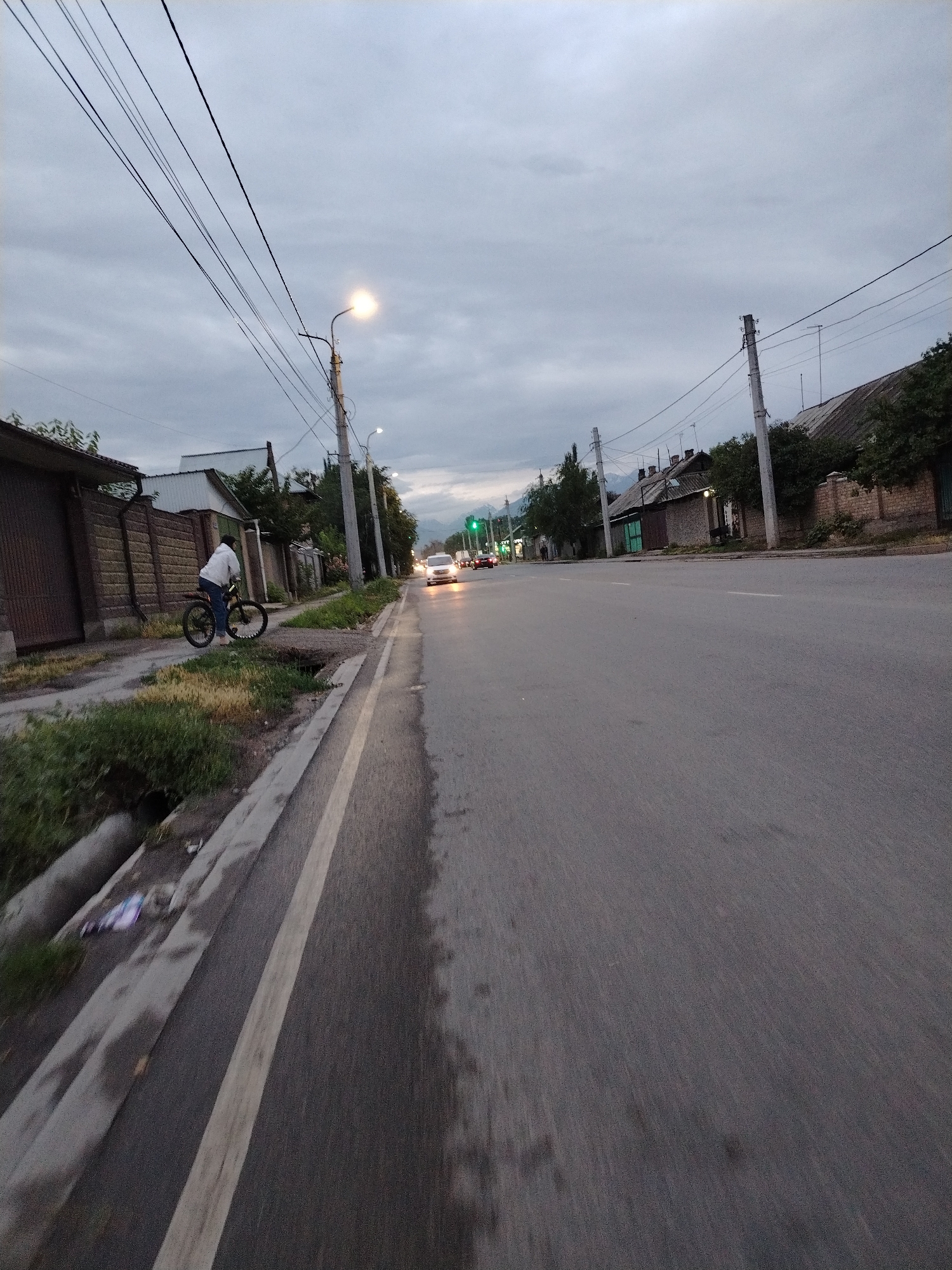 The image depicts a street scene in Bishkek City, Kyrgyzstan, during twilight. A young person on a bicycle is the primary subject, positioned on the right side of a paved road, facing away from the camera. The cyclist is wearing a white hooded jacket and blue jeans. The bike appears to be a mountain bike with knobby tires.
The setting is a residential street lined with houses and fences. On the left, a brick fence and a house are visible, with some greenery and a drainage ditch along the roadside. The road itself has a white lane marking and appears to be wet, suggesting recent rain or a damp day. A culvert is visible on the left, with some litter.
In the background, other cars are visible on the road, their headlights on, indicating low light conditions. Traffic lights, one showing green, can be seen in the distance. Utility poles with overhead wires stretch across the sky, which is overcast and grey, characteristic of dusk or a cloudy day. The overall atmosphere is subdued and calm, with a slight sense of motion blur, possibly from the perspective of someone riding a vehicle or walking quickly.
No visible text is present in the image. The time of day is late afternoon or early evening, judging by the lighting and the activated car headlights. The weather appears to be overcast and possibly humid. The location cues like the architecture of the houses and the utility poles are consistent with a Central Asian setting.