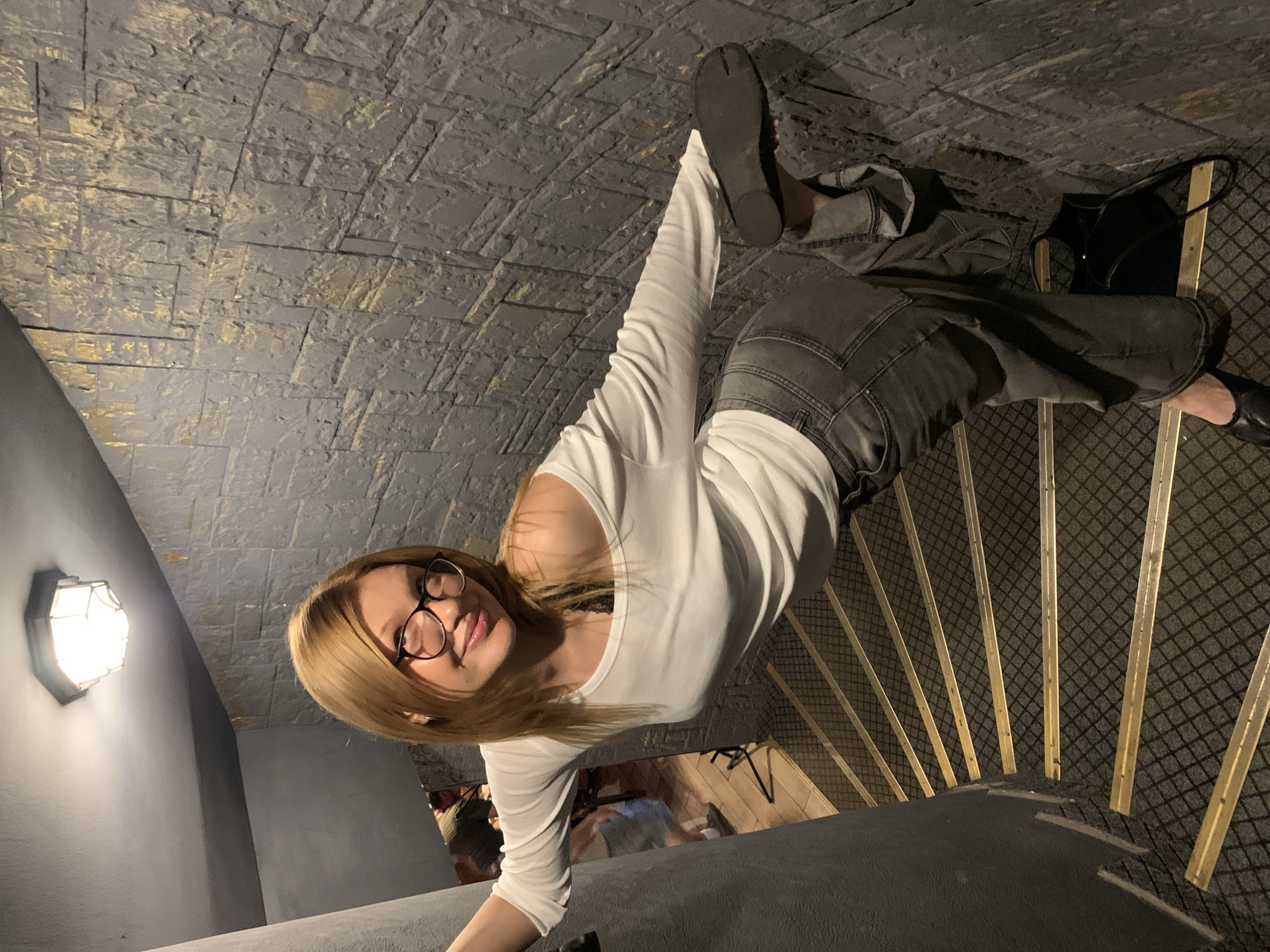 The image shows a young woman with blonde hair and glasses, wearing a white long-sleeved shirt and grey jeans. She is smiling and looking at the camera. She is standing on a staircase with grey stone walls and wooden steps. The staircase appears to be indoors, possibly in a bar or restaurant, given the presence of other people in the background. The lighting is dim, suggesting it is nighttime. The overall mood is casual and friendly.