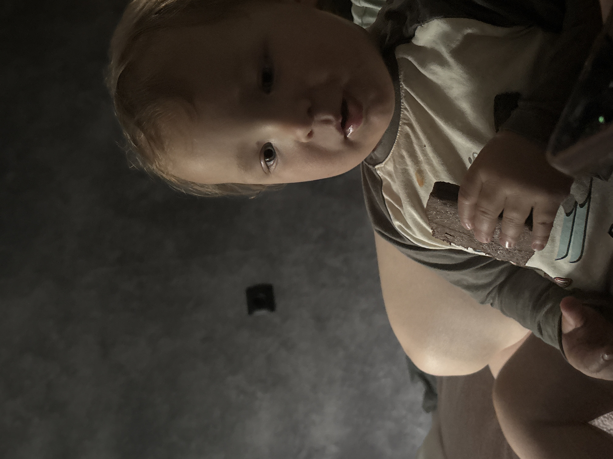 The image shows a close-up of a baby from a low angle, with the baby's face partially obscured by the camera's perspective. The baby has fair skin and light-colored hair. The baby is wearing a raglan-style shirt with a light-colored front and dark sleeves. The baby's mouth is open, and it appears to be holding a dark, rectangular object, possibly food, in its hand, bringing it towards its mouth. The background is a dark, textured surface, possibly a wall or floor. The lighting is dim, creating shadows and highlighting the baby's features. The overall impression is an intimate, candid moment captured at close range, focusing on the baby's interaction with the object. There is no visible text or discernible location cues.