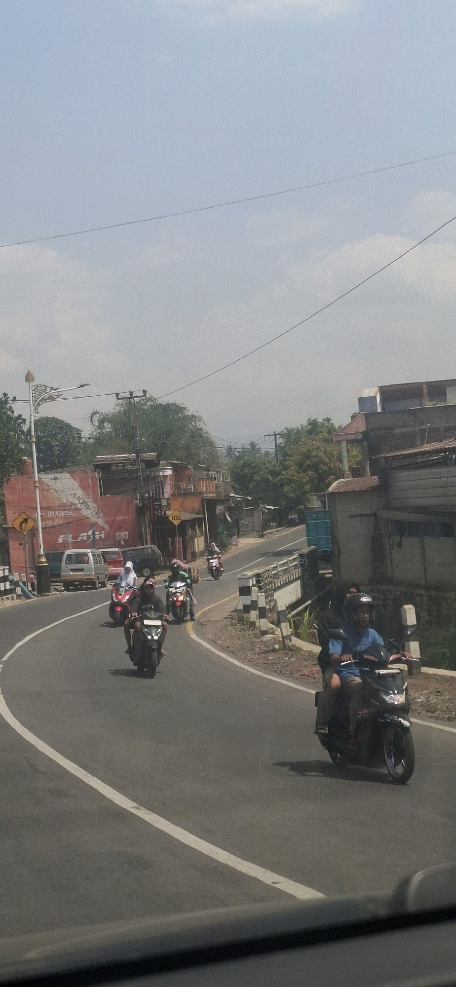The image captures a street scene in Cilaku, Indonesia, under a hazy, bright sky with a few wispy clouds. The road curves to the right, with a white line marking the edge of the lane. Several motorcycles are in motion. In the foreground on the right, a person wearing a blue shirt and a helmet rides a black motorcycle. Further ahead on the same lane, a group of motorcycles are traveling together. Two women, one in a red outfit and white headscarf and another in a green outfit and headscarf, ride separate motorcycles. A third motorcycle in this group is ridden by a person in a black hoodie. In the distance on the left side of the road, a small silver van is parked, and further back, a red building with white lettering that reads "TELKOMSEL FLASH" is visible. A decorative street lamp stands near the building. The background features more buildings, trees, and some hills in the distance. The environment appears to be a semi-urban or rural area with a mix of commercial and residential structures alongside the road.