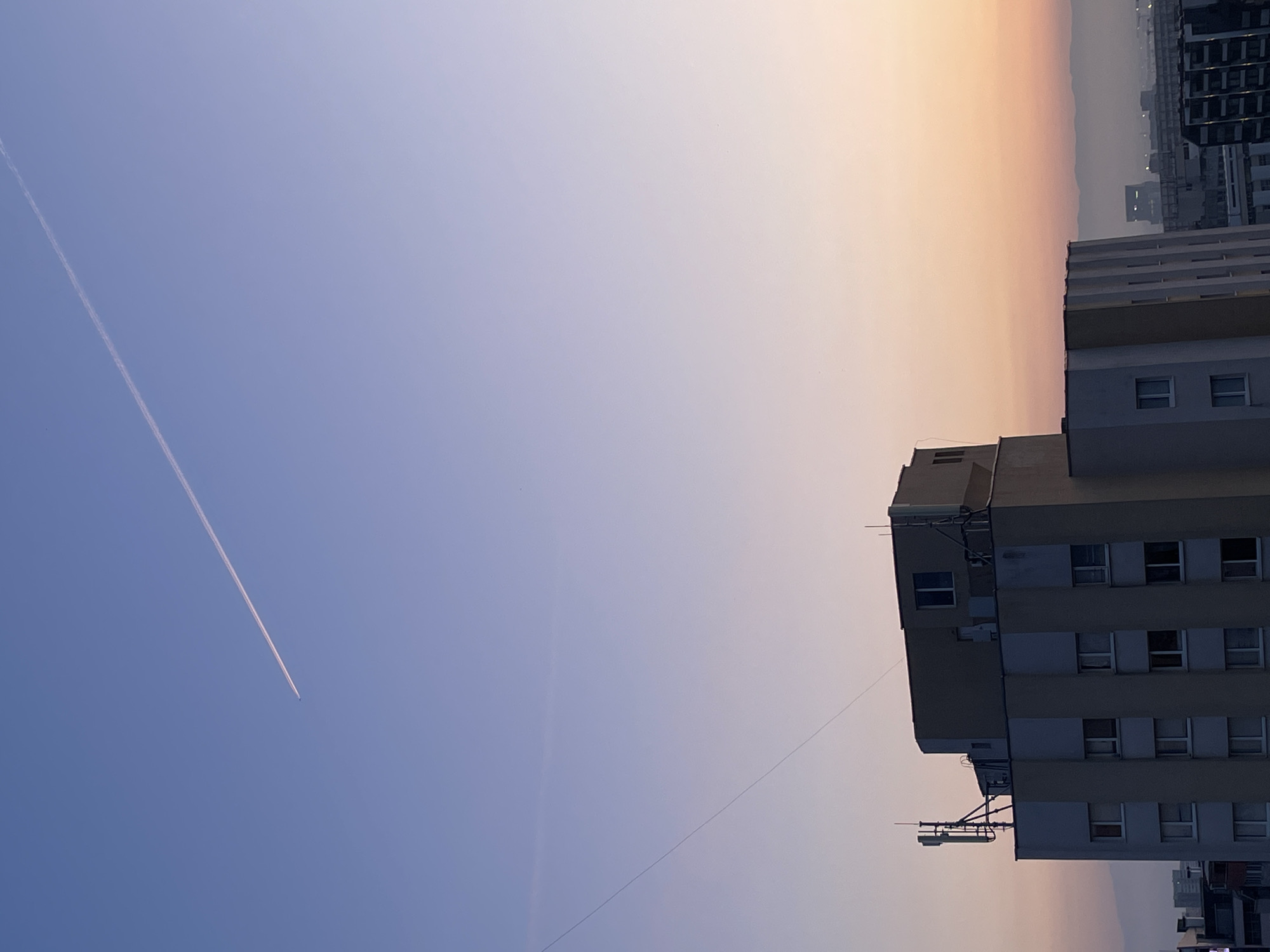 This image captures an urban landscape scene during what appears to be either sunrise or sunset, showcasing a striking sky gradient over buildings in Sofia, Bulgaria. The photograph is presented in a vertical orientation but depicts a horizontal view.
The upper and left areas of the frame are dominated by a clear sky, which transitions smoothly from a vivid, deep blue on the far left to a soft, warm orange-yellow towards the right. A prominent white contrail, left by an aircraft, stretches diagonally across the blue expanse of the sky. A small, dark speck, identified as an airplane, is visible at the trailing end of this contrail.
In the mid-ground and foreground, a multi-story building with a flat roof occupies the right side of the composition. Its facade is light beige or off-white, featuring multiple rows of rectangular, dark-paned windows. The roof and upper sections of the building are equipped with various antennas, satellite dishes, and other utility structures. A thin, dark wire or cable extends diagonally downwards from near the top of this building, crossing a portion of the sky.
Beyond the primary building, a hazy cityscape with additional structures stretches towards the distant horizon. These distant buildings are largely obscured by atmospheric haze, blending into the warm hues of the sky. The lighting suggests the "golden hour," providing soft, warm illumination. The weather appears clear, with only a slight haze visible in the distance. There is no discernible text present in the image.