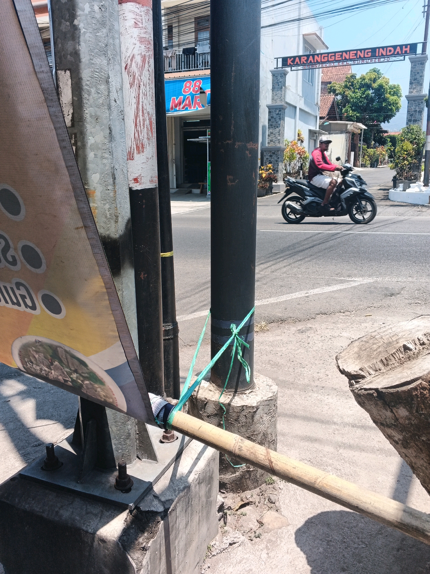 A motorcycle rider in a red shirt and white shorts passes under an archway in Semarang, Indonesia. The archway has the words "KARANGGENENG INDAH" in red LED lights. A banner with food imagery is partially visible in the foreground, held up by a bamboo pole. The scene is set on a sunny day, with clear blue skies. The road is paved, with a white line marking the lanes. In the background, there are buildings and trees. A chopped tree stump is visible on the right side of the image.