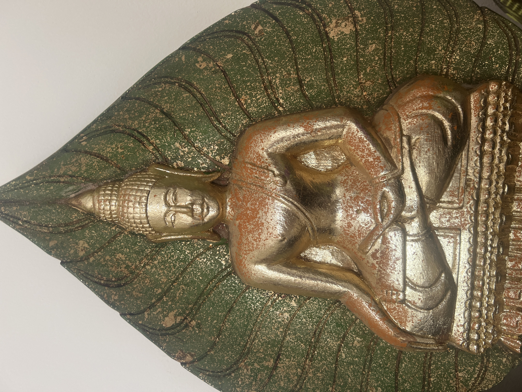 A close-up, head-on shot captures a golden statue of a seated Buddha. The Buddha is depicted with a serene expression, wearing traditional robes, and resting in a meditative pose. The statue is set against a backdrop of a large, green, leaf-shaped design, which appears to be part of the same artifact. The green leaf is textured and has visible lines suggesting veins. The gold of the Buddha statue exhibits an aged patina, with some areas showing a reddish-brown undertone, suggesting wear or intentional decorative detail. The lighting is even, highlighting the contours of the statue. The background wall is plain and white. The image composition focuses on the upper body and seated posture of the Buddha, with the leaf-shaped element framing it from behind. No specific location in Ko Samui District, Thailand can be determined from the visual information provided.
