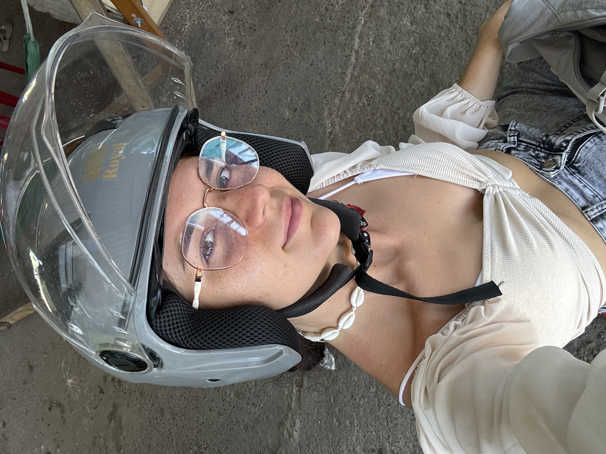 A young woman wearing a gray helmet with a clear visor and gold "Royal" lettering, rose-tinted round glasses, and a white seashell necklace is pictured. She has a fair complexion with freckles across her nose and cheeks. She is wearing a light cream-colored tied-front top over a white bikini top, and distressed denim jeans. The background appears to be an outdoor area with a concrete or asphalt ground. The lighting suggests it is daytime, possibly outdoors in a humid climate, consistent with the city of Hong Son, Vietnam.