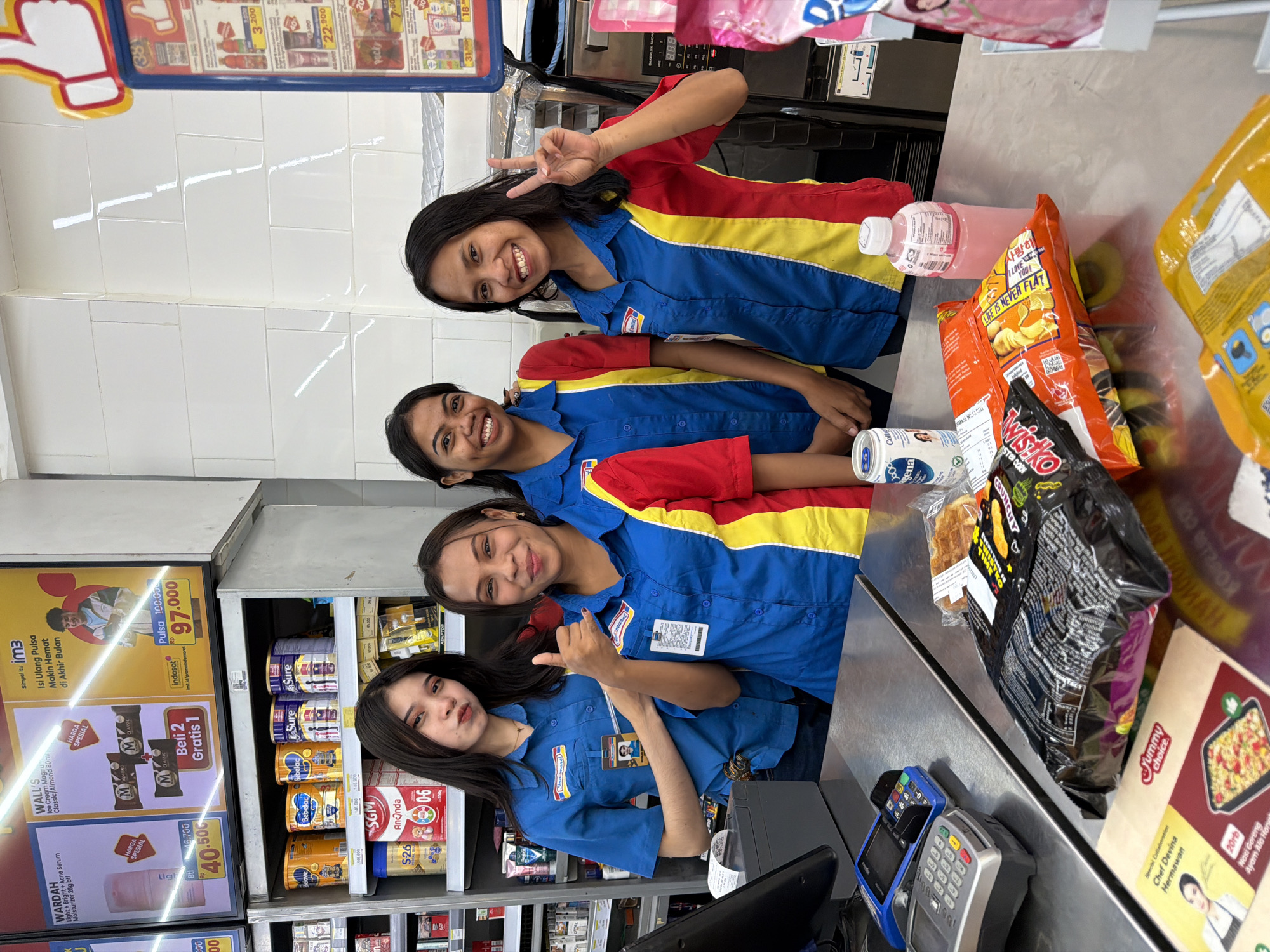 This is a candid, eye-level photograph taken indoors, likely in a convenience store in Kuta, Indonesia, given the visible signage and products. The scene is a checkout counter, with three young women, presumably employees, posing for the camera.
The two women in the foreground are wearing matching blue shirts with red and yellow accents and company identification badges. They are smiling and appear cheerful and friendly. The woman on the left is making a peace sign with her fingers, while the woman to her right gives a thumbs-up. The third woman is partially visible in the background, also smiling and wearing a similar uniform.
On the counter, various products are displayed, including bags of snacks, drinks in bottles and cans, and what appears to be a payment terminal. Behind the counter, shelves are stocked with food items, including canned milk, juice, and other packaged goods. Advertisements are also visible, promoting mobile phone top-ups and ice cream. The overall lighting suggests it's daytime, and the atmosphere is lively and welcoming. The visible text includes product names, prices, promotions, and brand logos, with a mix of Indonesian and English.