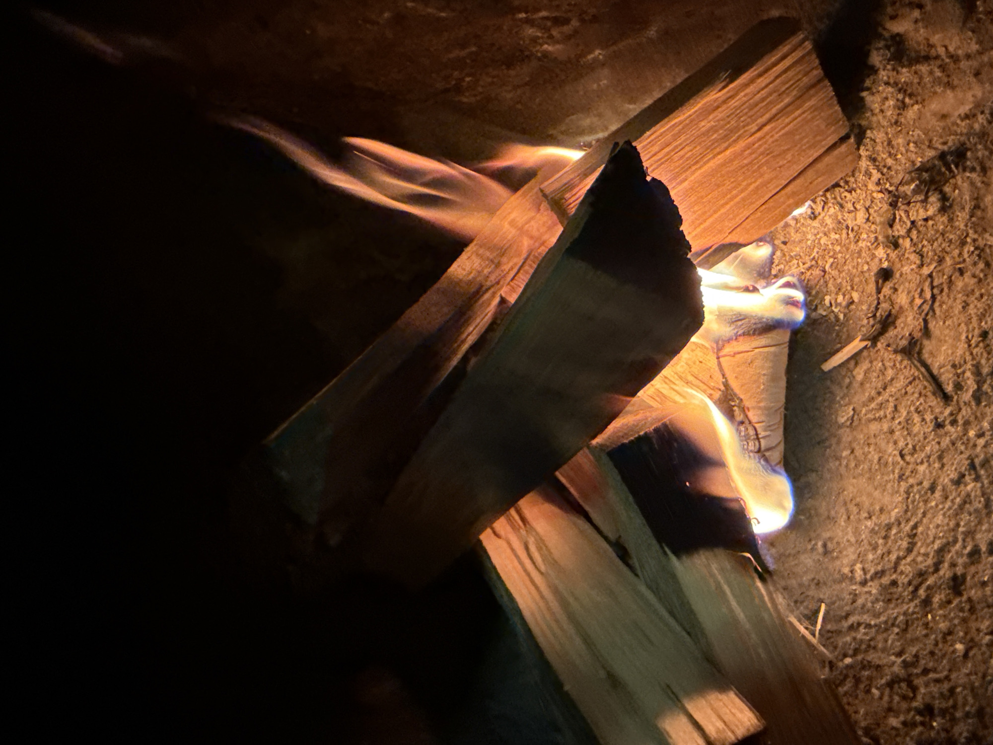 The image shows a close-up view of a bonfire burning in an outdoor setting, possibly a fireplace or fire pit, in Нагаево, Russia. The fire is actively consuming a stack of dry wooden logs, which are arranged in a haphazard manner. Flames, ranging from yellow to orange and with hints of blue at the base, are visible licking the wood. The surrounding environment appears to be dark, with the light from the fire illuminating the immediate area. The ground beneath the fire is covered with ash and debris. The scene is dark, with the flames being the primary source of light.