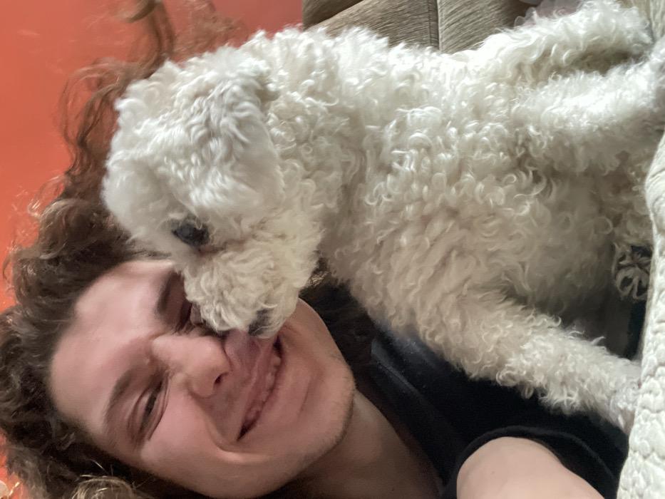 The image depicts an indoor scene focusing on the close interaction between one human male and one dog. A male individual, appearing young adult with fair skin and long, wavy brown hair, occupies the left side of the frame. He is positioned at an angle, looking towards the right, with a wide smile that partially closes his eyes and reveals his teeth. His tongue is slightly extended. He wears a dark-colored garment.
To his right, occupying the central and right portions of the frame, is a small, white, curly-haired dog, resembling a Bichon Frise or Poodle type. The dog's head is turned towards the man's face, and it is actively licking the area around the man's mouth. The dog is seated upright on a light-colored, textured surface, possibly a quilted blanket or bedspread.
The background consists of a plain, warm orange-colored wall behind the subjects. In the upper right corner, a segment of a brown, possibly upholstered or wooden, object is visible, suggesting furniture. The overall environment appears to be a domestic setting. No visual information within the image frame identifies the location as the city of Yekaterinburg, Russia.