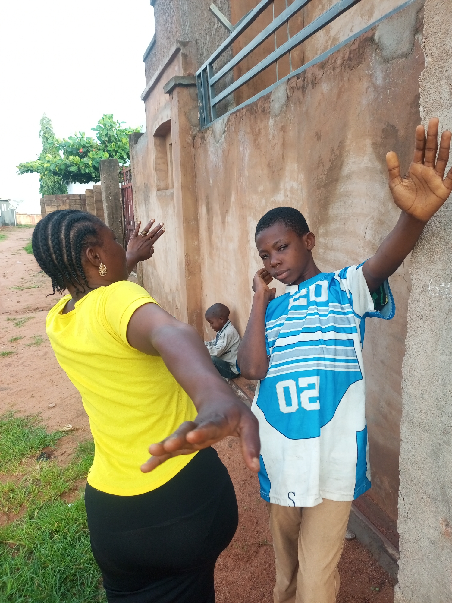 In Tempure, Nigeria, a woman with braided hair, wearing a yellow t-shirt and black pants, stands in the foreground with her back to the camera, extending her right arm outwards as if reaching or gesturing. Beside her, a young boy in a blue and white striped t-shirt with the number "02" prominently displayed, leans against a textured, light brown wall. His left arm is raised, and his hand is near his ear, while his right hand rests on the wall. In the background, another child, partially obscured, sits near the wall. The environment appears to be an outdoor urban setting with a dirt and grassy ground, a concrete wall with a metal grate at the top, and some greenery and trees visible to the left.