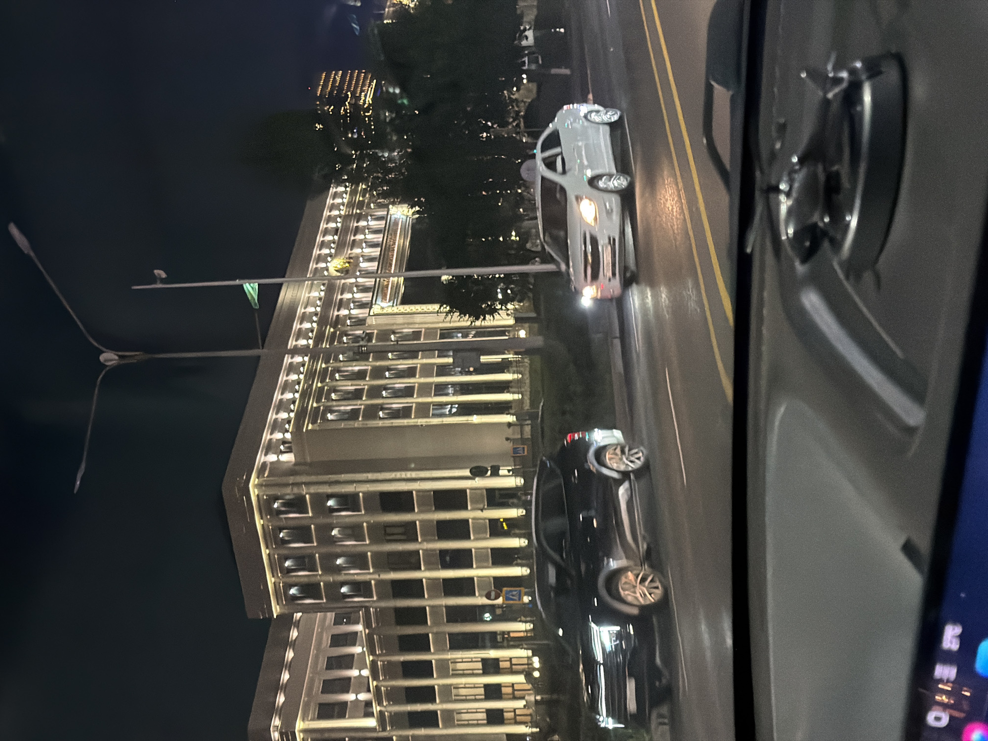 A nighttime street scene in Astana, Kazakhstan, shows a modern, illuminated building with multiple floors and a symmetrical facade, featuring rows of windows and vertical accents. In the foreground, a dark SUV drives on the right side of the road, followed by a white sedan. The street is wet, reflecting the lights from the building and streetlights. The view is from inside a vehicle, looking over the dashboard where a small, silver helicopter figurine is visible. The sky is dark, with some lights from a distant building visible.