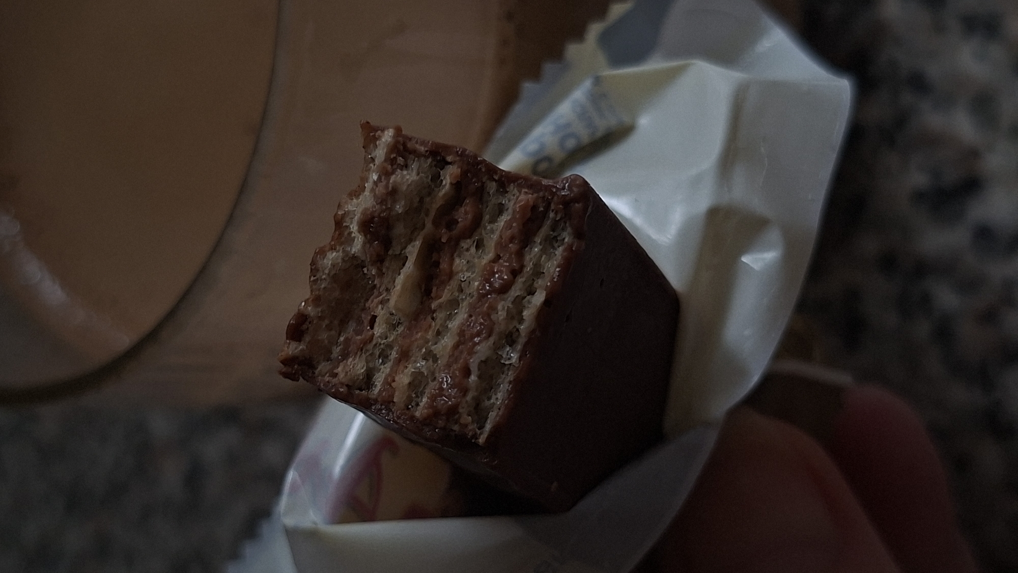 A close-up, slightly blurred, low-angle shot shows a bite taken out of a rectangular chocolate-covered wafer snack. The snack is mostly brown with visible layers of wafer and a creamy chocolate filling. It rests inside its white, crinkled paper wrapper, which is slightly torn. In the background, a light brown liquid, possibly coffee or hot chocolate, fills a glass. The surface beneath the snack and glass appears to be a speckled countertop. There are no discernible people, or specific landmarks that would indicate a location in Myadzyel, Belarus.
