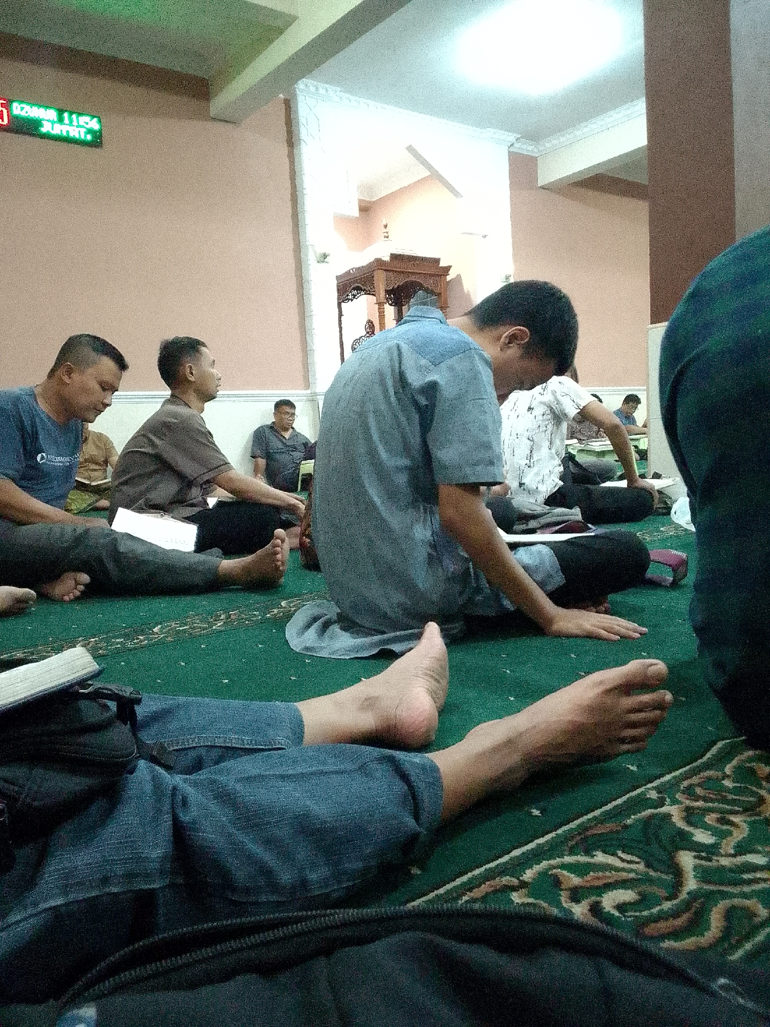 The image depicts an indoor scene, likely a mosque, with several individuals seated on a green carpeted floor. In the upper left corner, a digital clock displays the time "11:54" and the day "JUMAT" (Friday), suggesting a Friday prayer time in Kertajaya, Indonesia.
The foreground shows the lower legs and feet of at least two individuals dressed in denim jeans. One person's bare foot is prominently visible, resting on the carpet. In the middle ground, several men are seated, some with books or papers in front of them. One man in a blue shirt is on the left, facing away from the camera, while another in a brown shirt is to his right. Further back, more men are seated, including one wearing glasses and a dark shirt. The background features a pinkish wall and a wooden architectural element, possibly part of a minbar or decorative structure. A white ceiling with lighting is visible at the top. The overall atmosphere appears to be one of communal gathering or prayer.