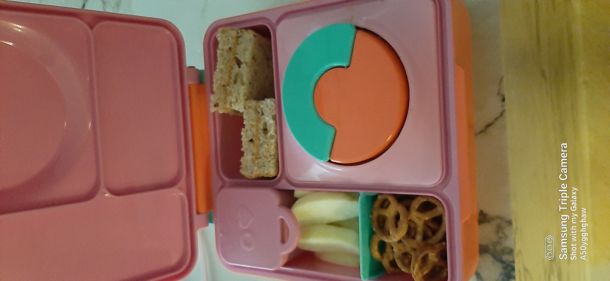A top-down medium shot captures a pink bento box filled with food items. The bento box has multiple compartments. One compartment holds two halves of a sandwich. Another compartment contains sliced apples and pretzels. A circular compartment is partially filled with an orange disc topped with a teal curved piece. The bento box is placed on a light-colored surface with a subtle marble pattern. The bottom of the image displays a watermark indicating "Samsung Triple Camera Shot with my Galaxy A50yyghghaw." The scene is indoors and appears to be a domestic setting, possibly a kitchen counter. There is no identifiable location in Dubai, United Arab Emirates.