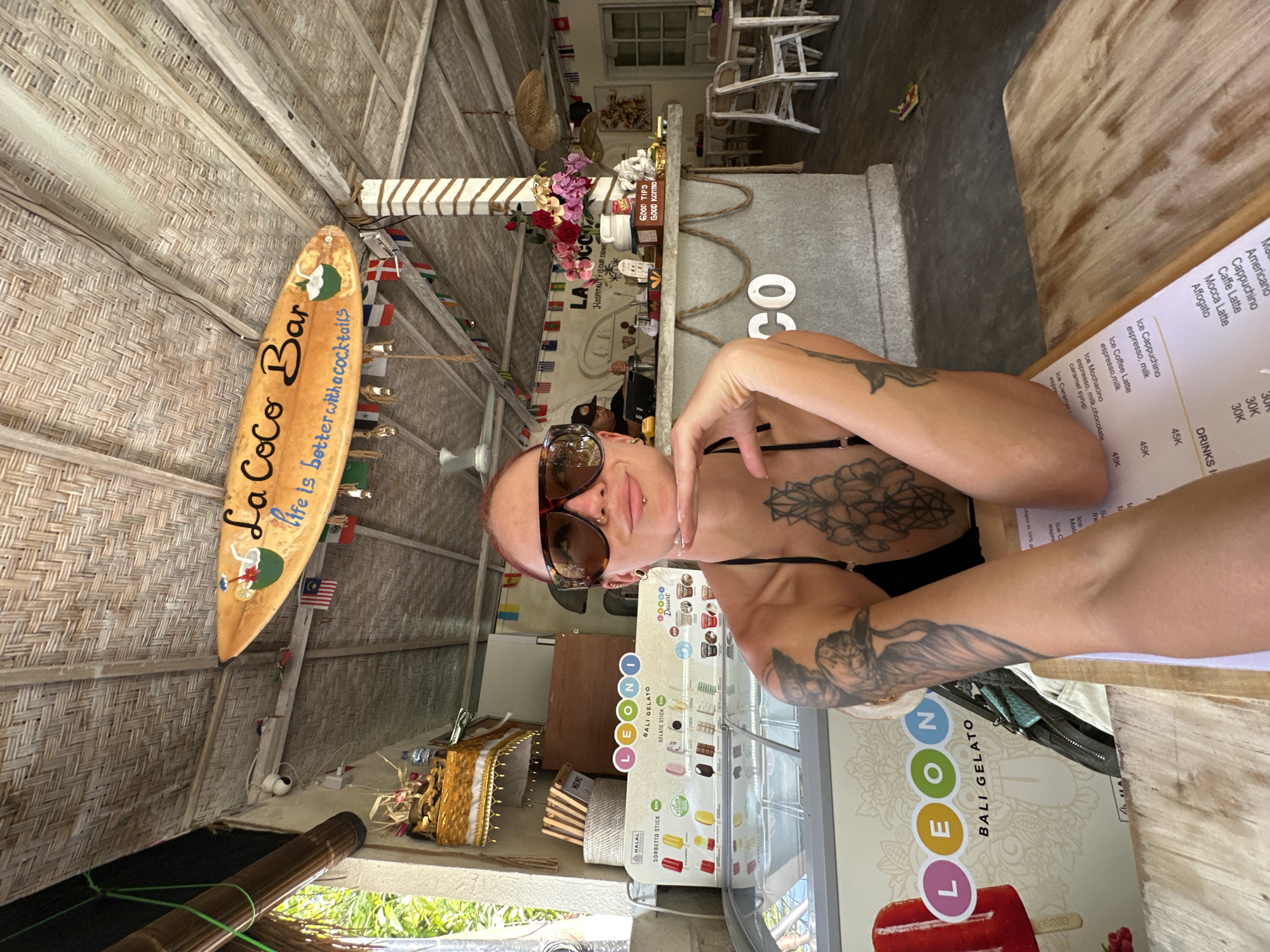 The photograph captures an individual with tattoos and sunglasses, seated at a table under a thatched roof in Nusa Penida, Indonesia. In the background, a surfboard sign reading "La Coco Bar life is better with a cocktails", along with a gelato display and a menu are visible. A bar area decorated with flags is also visible.