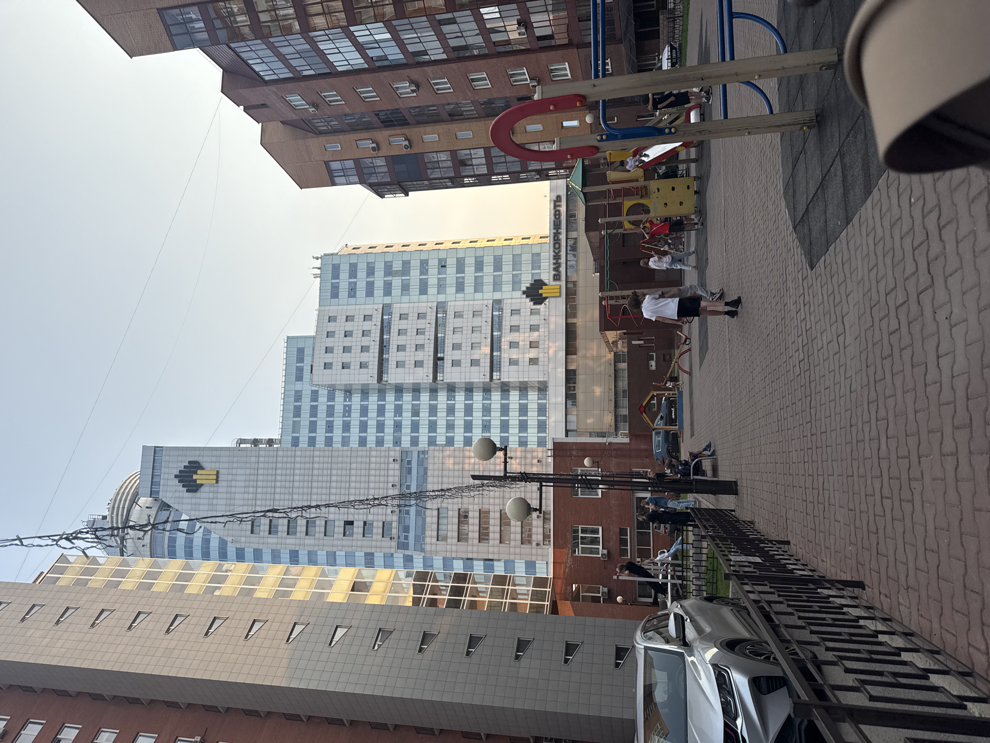 The image depicts a courtyard in the city of Krasnoyarsk, Russia, with a playground and residential buildings. A silver Geely Coolray car is parked on the left side of the frame. The foreground shows a paved courtyard with a black metal fence. In the mid-ground, a playground with slides, swings, and climbing structures is visible, with several children playing. Behind the playground are low-rise brick buildings. In the background, tall modern residential buildings dominate the skyline, with one prominent building featuring the logo "BANKOONEOFT." The sky is clear and a pale blue, suggesting it is daytime.