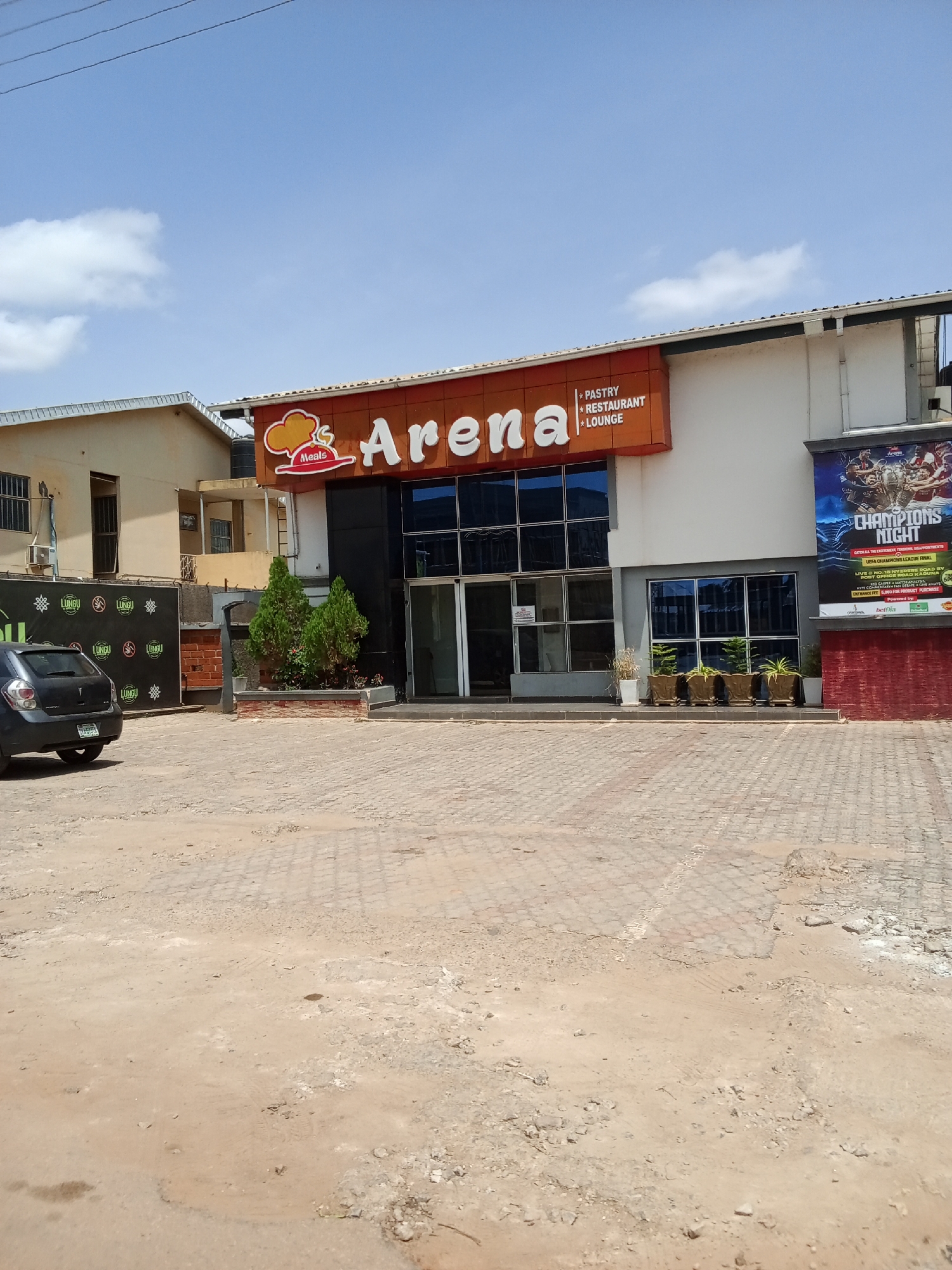 The image shows the exterior of a building that houses "Arena" which is a pastry, restaurant, and lounge. The building has large glass windows and a prominent orange sign with the "Arena" logo. To the right of the entrance, a large poster advertises a "Champions Night" event for the "UEFA Champions League Final". A black car is parked in front of the building on a paved area. The surrounding environment appears to be a street with a few trees and a partially paved ground. The sky is blue with some clouds. The building is situated in Narayi, Nigeria.