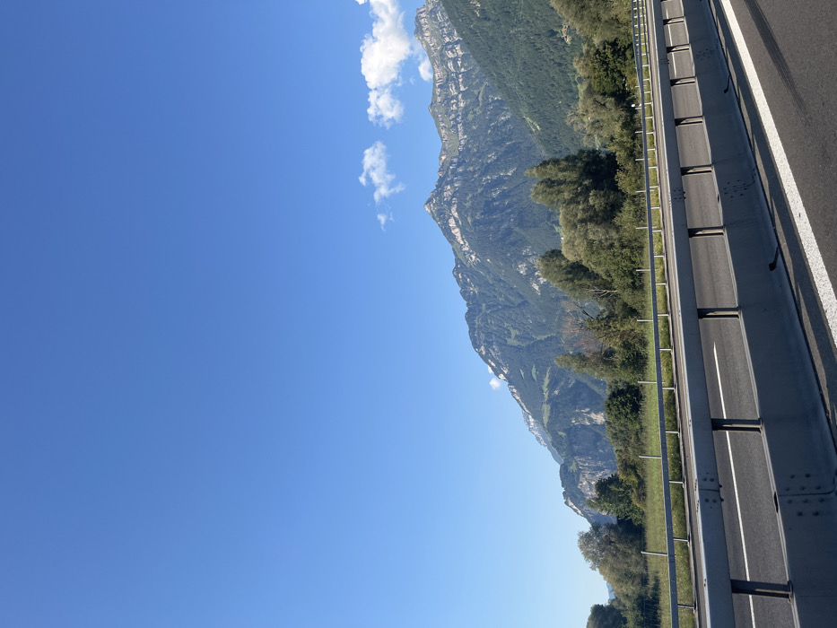 The scene is a daytime exterior view of a highway section situated in Seedorf, Uri canton, Switzerland. The lower portion of the image displays an asphalt roadway with a prominent solid white line marking its edge. Adjacent to the roadway, a grey metal guardrail assembly, supported by vertical posts anchored into a concrete base, extends horizontally across the frame. Beyond the guardrail, a flat, verdant expanse is visible, featuring scattered mature trees with full green foliage and a thin, horizontal fence line. This area transitions into dense green tree cover that ascends the lower and mid-slopes of a substantial mountain range. The background is dominated by towering, rugged mountains, distinguished by extensive green vegetation on their lower sections and exposed grey rock formations with distinct horizontal strata at higher elevations, culminating in sharp peaks. The sky above is clear and bright blue, with a few small, scattered white cumulus clouds visible above the mountain ridges on the right side of the frame. No individuals, vehicles, or animals are present within the view.