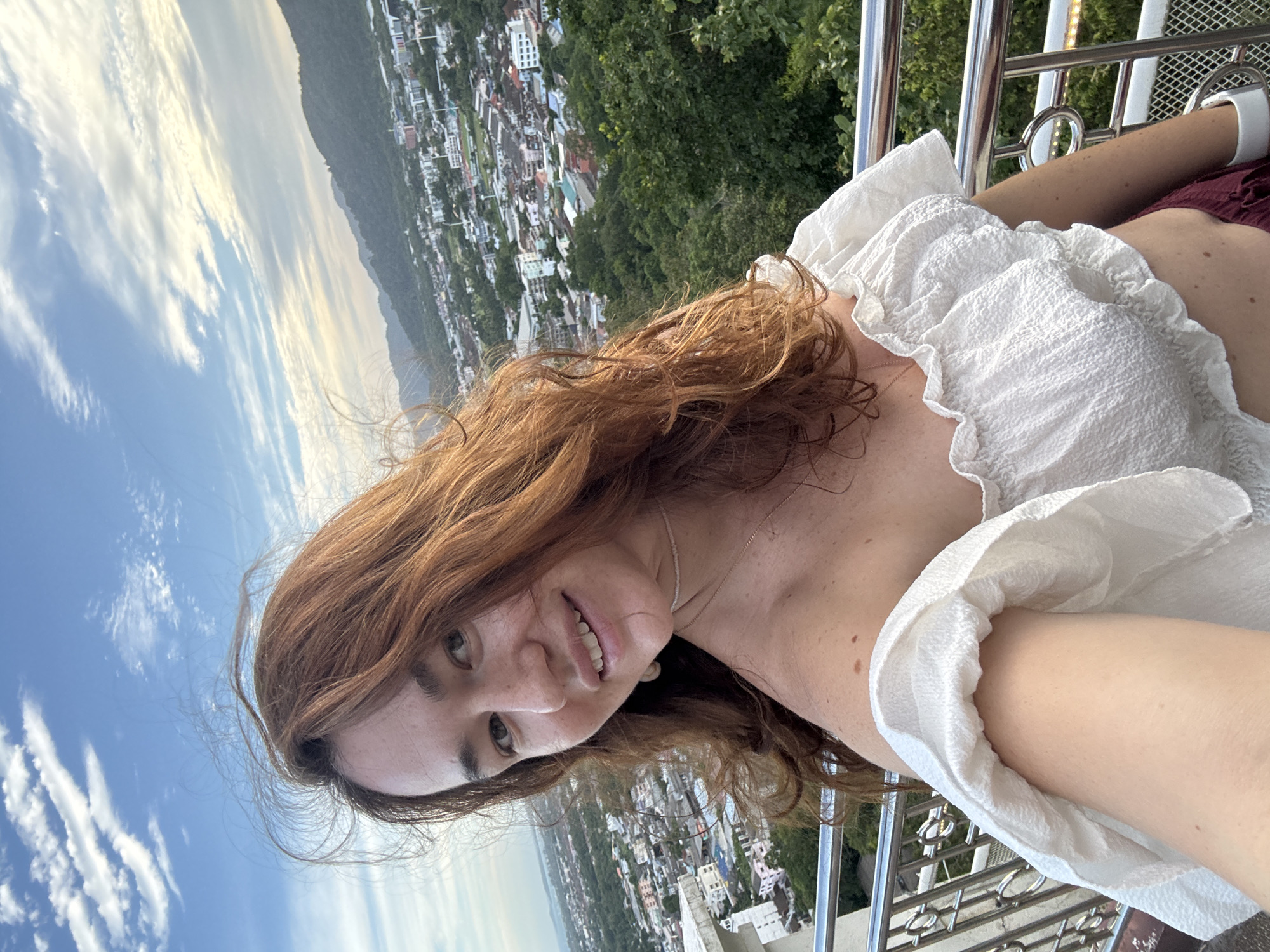 A medium shot captures a young woman with long, reddish-brown hair and fair skin, taking a selfie from a viewpoint overlooking a cityscape. She is wearing a white, off-the-shoulder crop top with ruffled trim. Her head is tilted slightly to her right, and she is smiling at the camera. In the background, a sprawling city with buildings and lush green hills can be seen under a partly cloudy sky. The railing of the observation deck she is standing on is visible in the foreground. This vantage point appears to be from a higher elevation, offering a panoramic view of the urban and natural landscape of Ratsada, Thailand.