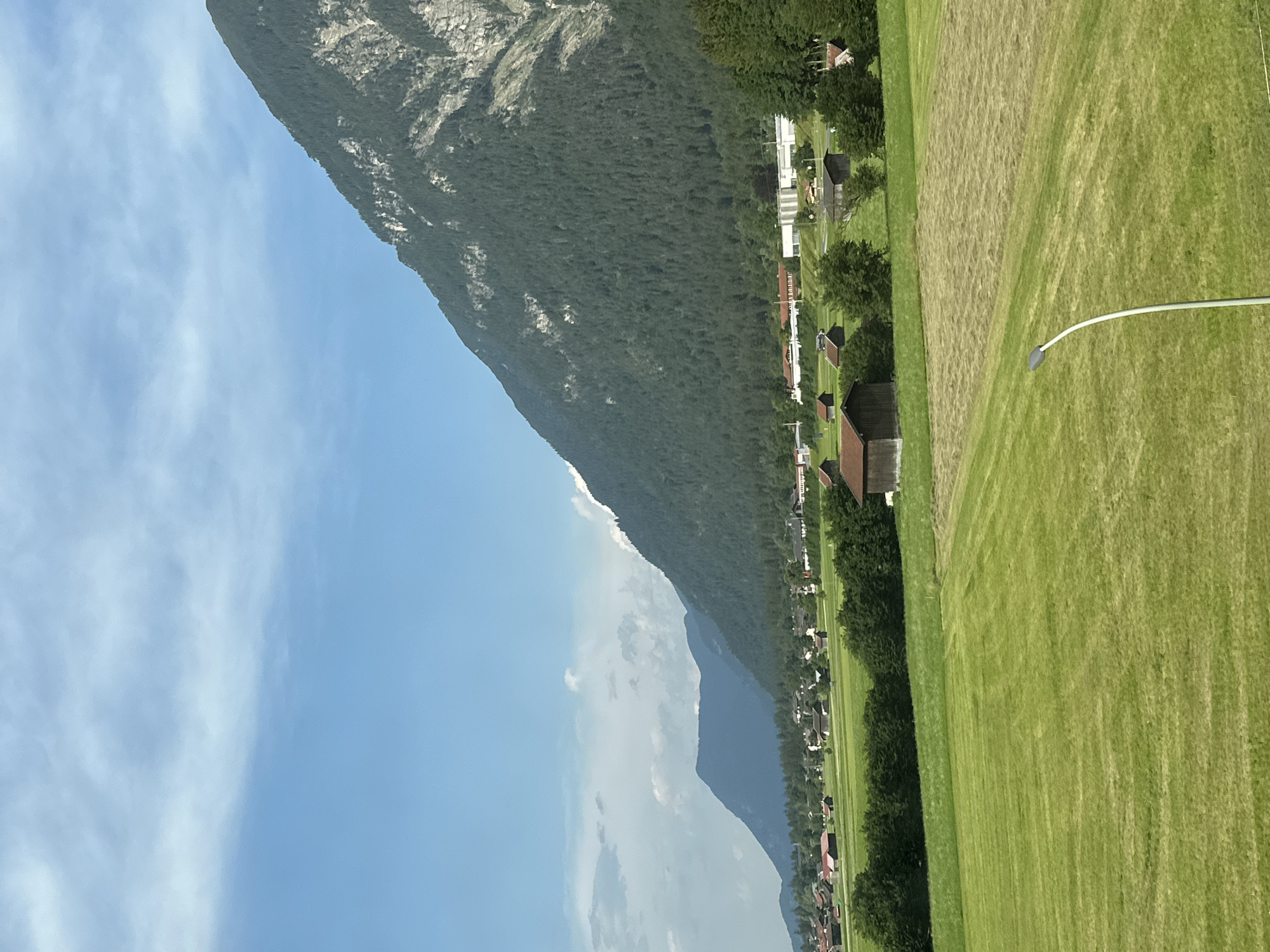 The image depicts a scenic rural landscape with a prominent mountain range in the background. In the foreground, there are expansive green fields, some of which have been mowed, creating a patchwork effect. Scattered across the fields are several small, rustic buildings with dark roofs, resembling barns or sheds. A village or small town is visible in the middle ground, characterized by houses with red and white roofs, nestled amongst trees. Further in the distance, more houses and structures, possibly industrial buildings, are present. The mountain itself is covered in dense green forest, with rocky outcrops visible higher up. The sky is a clear blue with some scattered white clouds. This scene is consistent with the geographical features found near Garmisch-Partenkirchen, Germany, known for its Alpine scenery and surrounding villages. There are no people or animals visible in the image. A solitary, modern street lamp stands in the right foreground, its grey pole curving upwards.